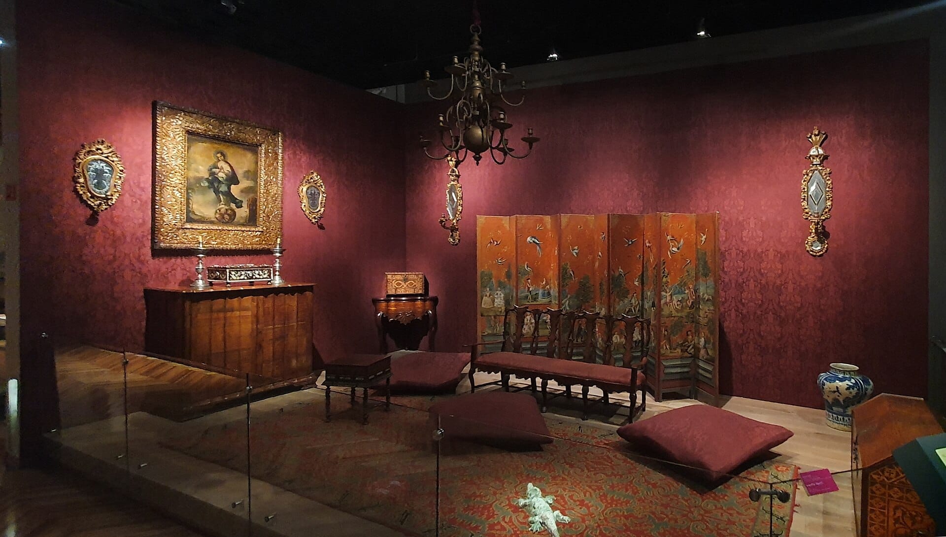 Esposizione al Museo Franz Mayer con elaborati mobili coloniali, opere d'arte religiosa con cornici dorate, paravento decorativo e cuscini morbidi in una sala dall'illuminazione calda.