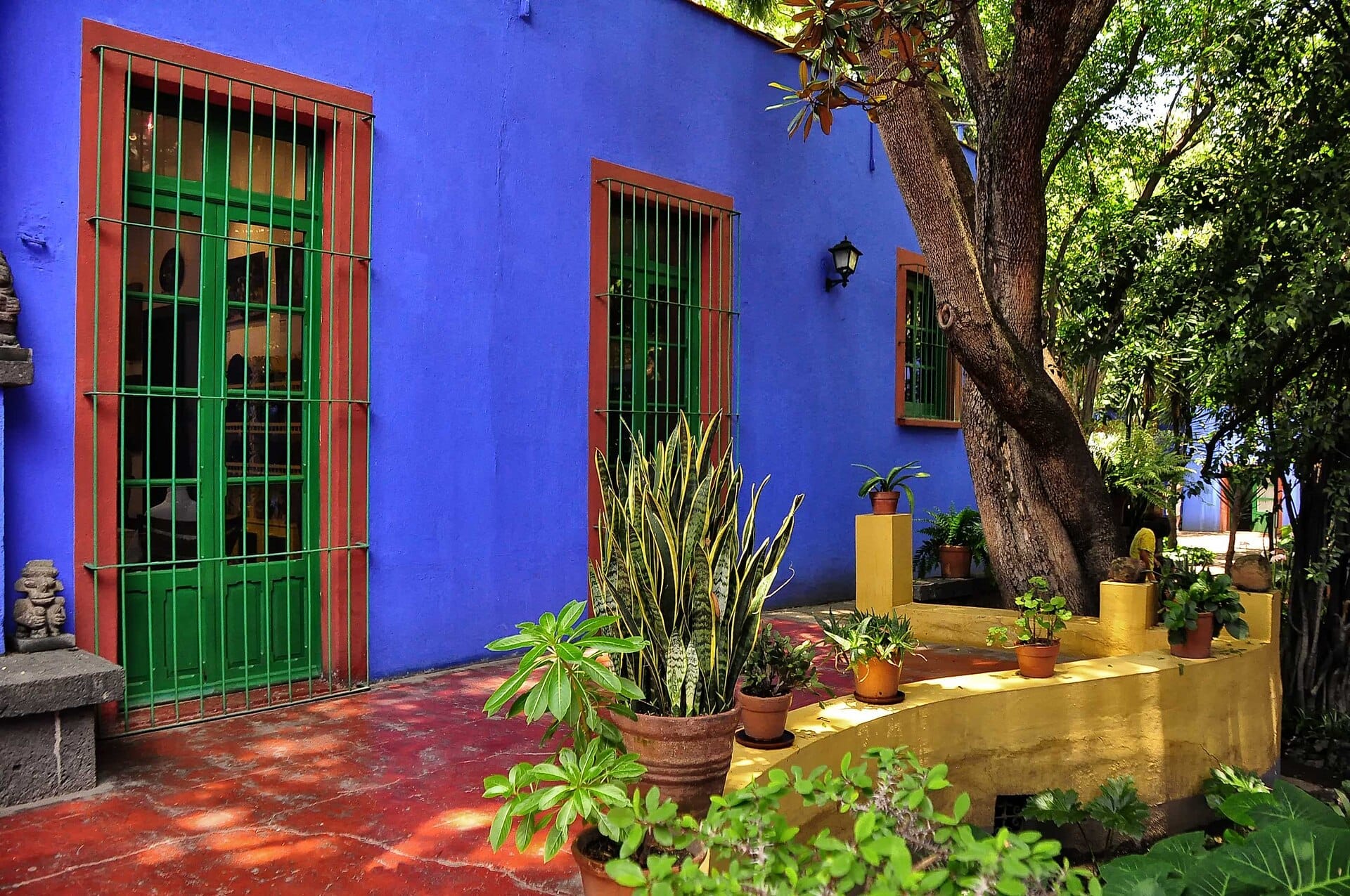 La vivace facciata blu del Museo Frida Kahlo, con finestre a grate verdi, piante in vaso, alberi ombrosi e luce solare screziata nel cortile.