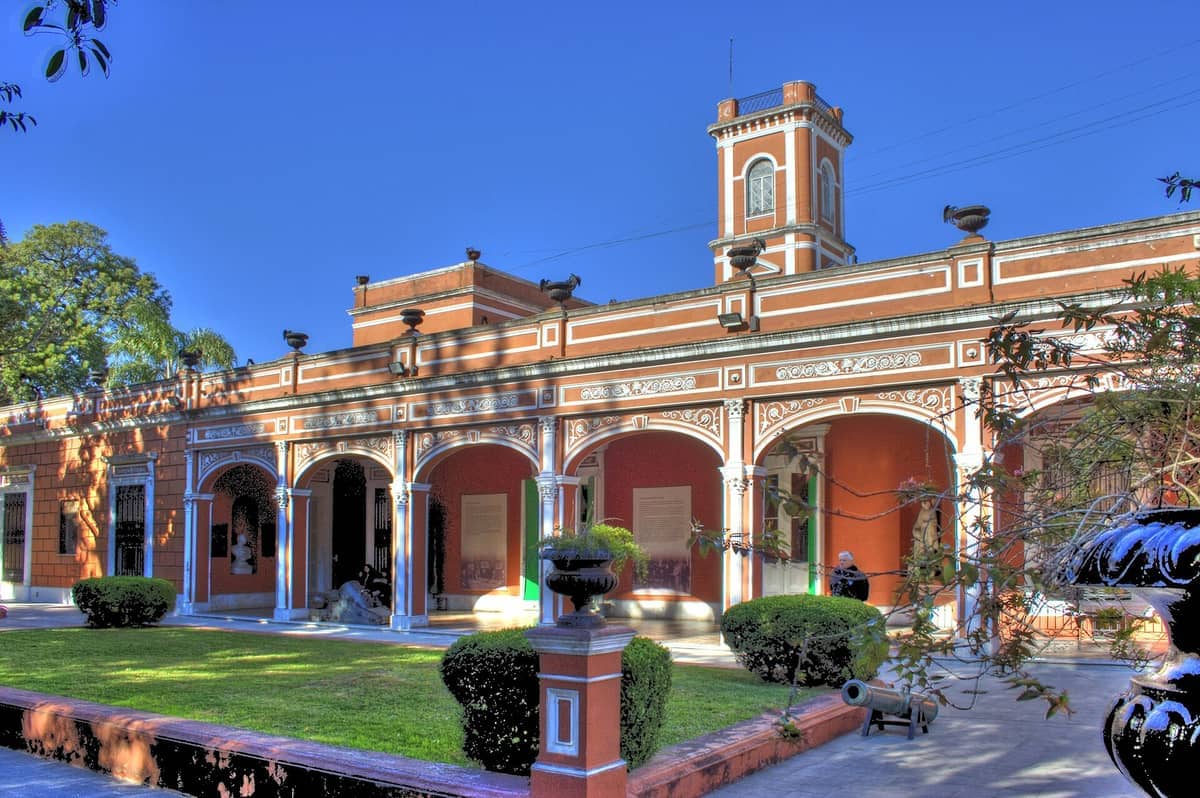 มุมมองด้านหน้าของ Museo Histórico Nacional พร้อมด้านหน้าอาคารสไตล์โคโลเนียลสีแดงและขาว ทางเข้าโค้งอาร์ช สวนที่ดูแลอย่างดี และท้องฟ้าสีฟ้าแจ่มใส