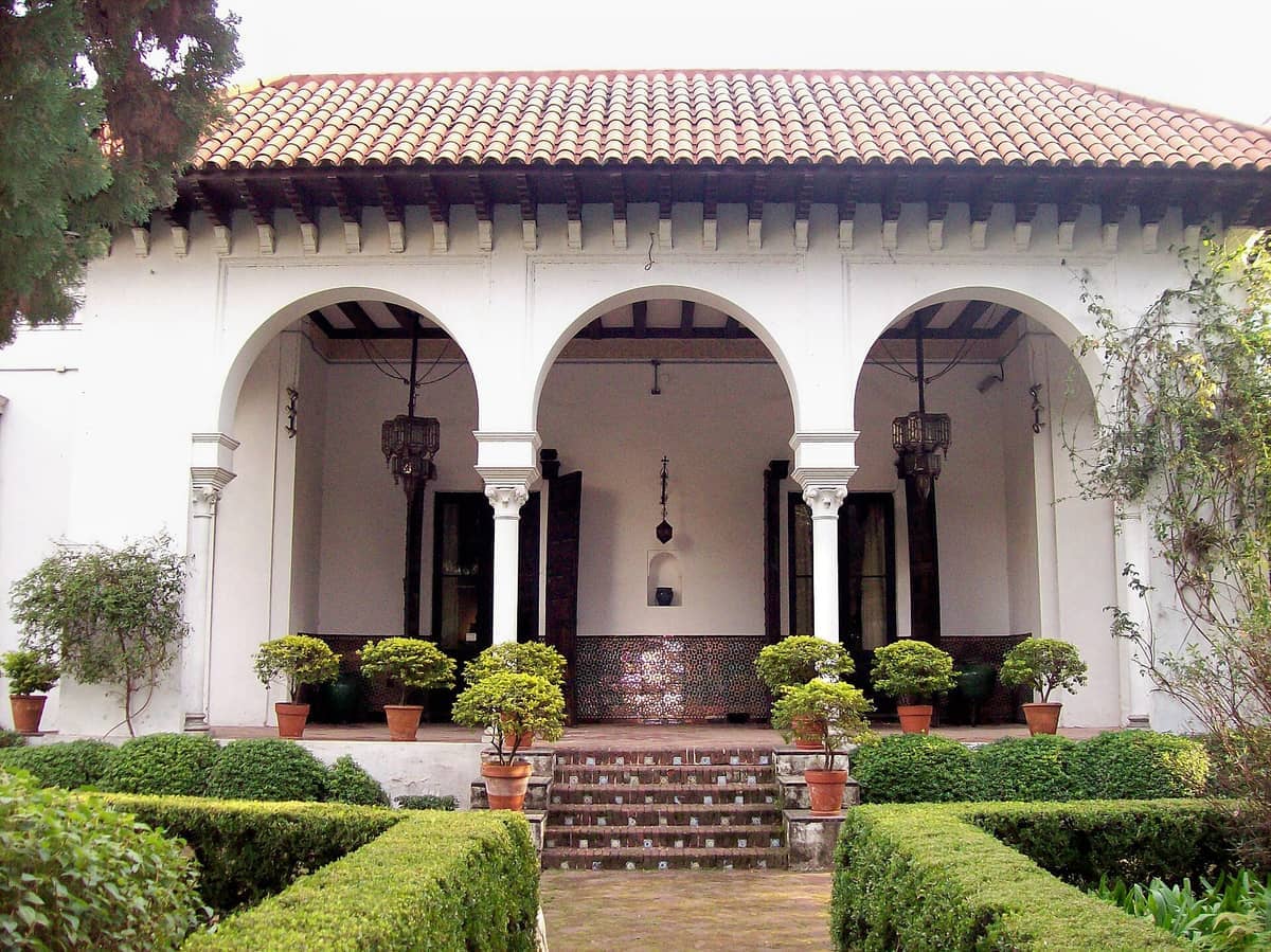 มุมมองด้านหน้าของ Museo de Arte Español Enrique Larreta ที่โดดเด่นด้วยซุ้มโค้งสีขาว หลังคาดินเผา ต้นไม้กระถาง และพุ่มไม้ตัดแต่งสไตล์สวนอันดาลูเซีย