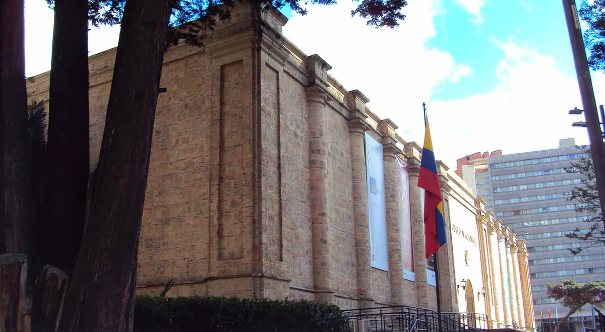 Exterior view of the Museo Nacional de Colombia, showing its stone facade, banners, Colombian flag, and part of the Centro Internacional skyline.
