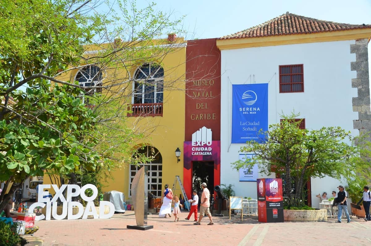 Facade of Museo Naval del Caribe in Cartagena with colorful colonial architecture, large trees, event banners and visitors walking past in the sunny plaza.