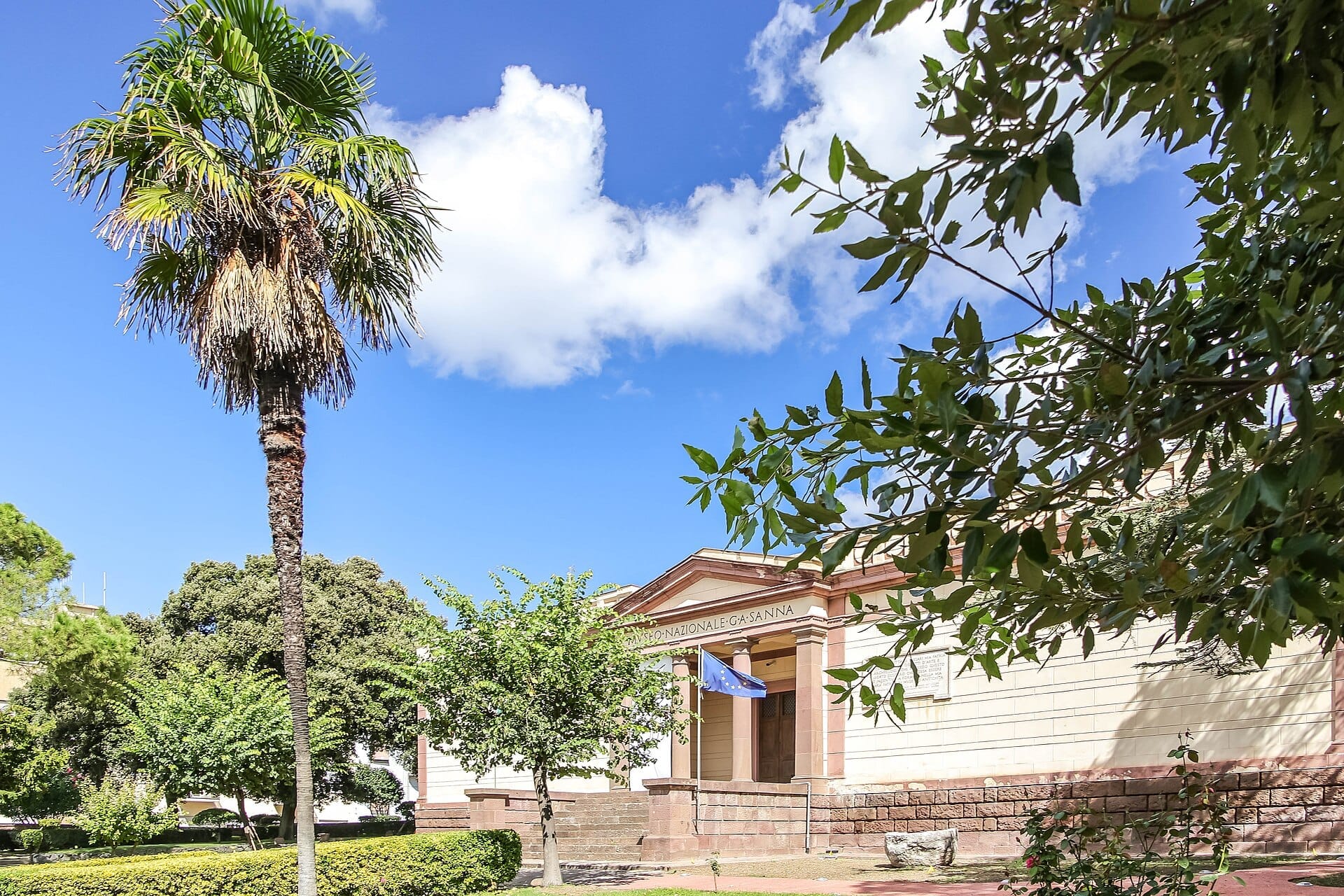 Das Museo Nazionale G.A. Sanna in Sassari, Sardinien, ist hinter üppigen Gärten, einer hohen Palme und unter einem strahlend blauen Himmel zu sehen.