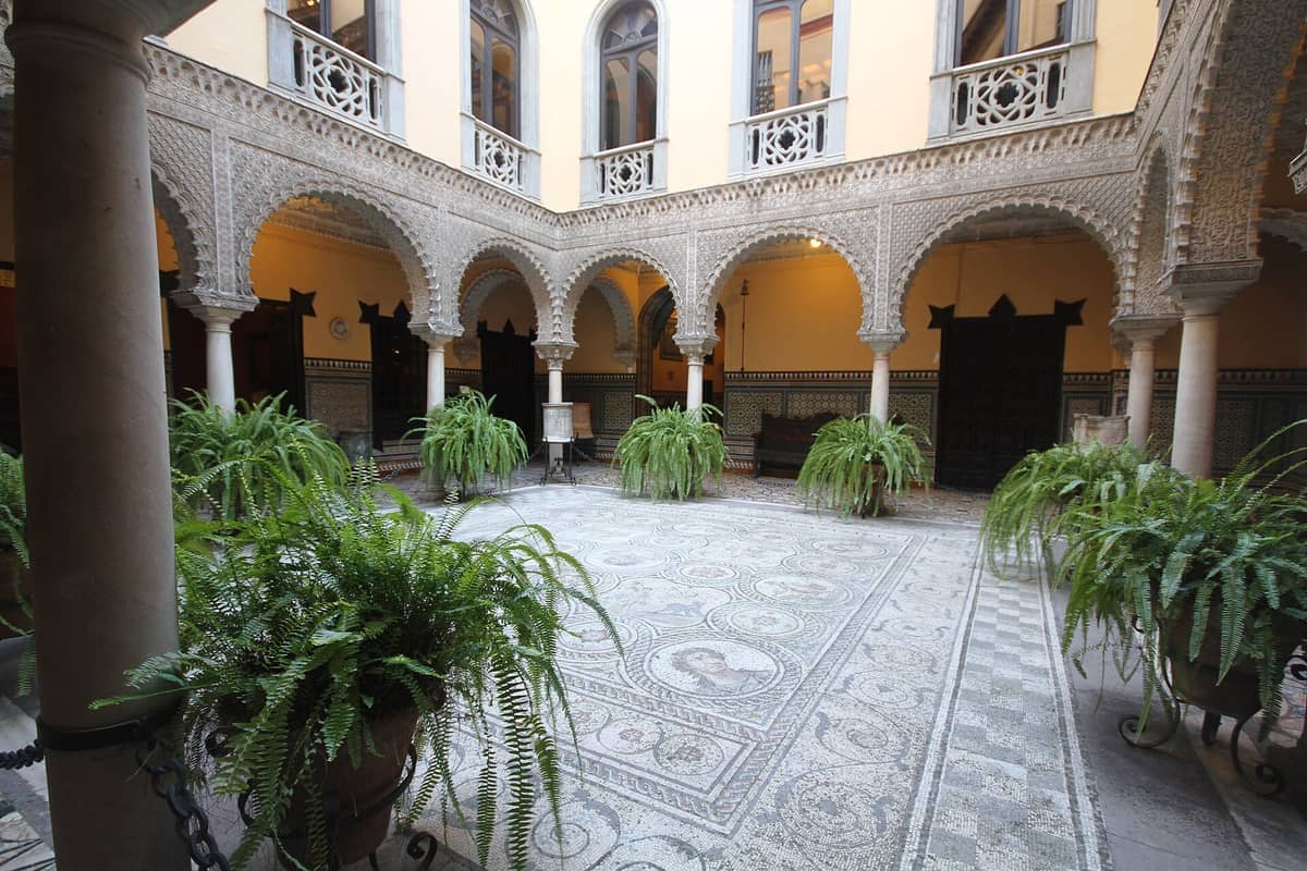 ลานภายในของ Museo Palacio de la Condesa de Lebrija ที่มีพื้นโมเสกโรมัน เฟิร์นสีเขียวชอุ่ม และซุ้มโค้งแกะสลักอย่างประณีตในเมือง Seville