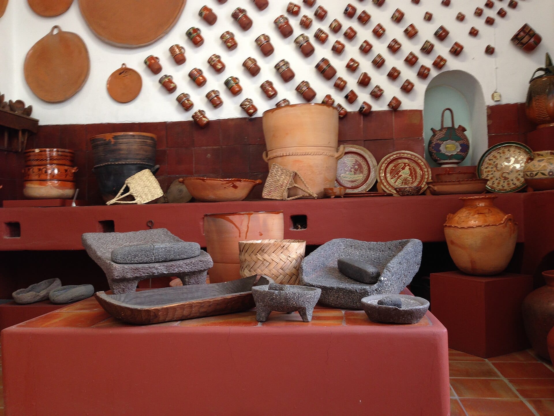 Vasi in ceramica tradizionali, piatti e macinatori di pietra sono esposti contro una parete rossa e bianca al Museo Regional de la Cerámica de Tlaquepaque.