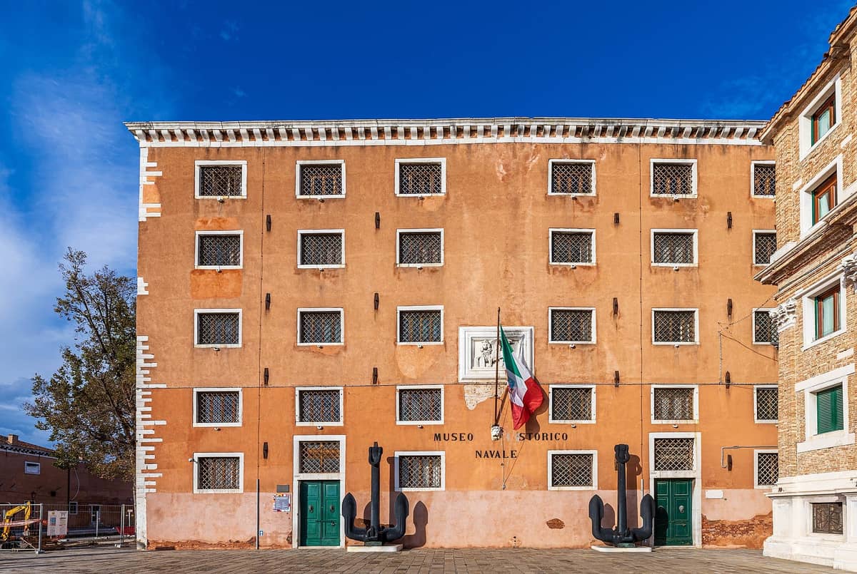 Frontfassade des Museo Storico Navale in Venedig – ein historisches ockergelbes Gebäude mit grünen Türen, vergitterten Fenstern, großen Ankern und der italienischen Flagge vor blauem Himmel.