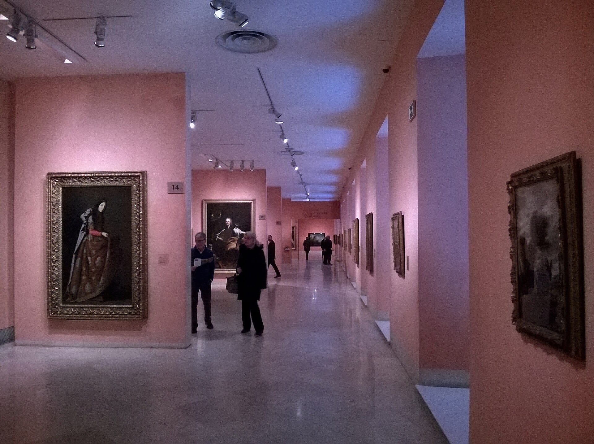 Galleria interna del Museo Thyssen-Bornemisza con dipinti incorniciati, pareti rosa tenue, illuminazione a spot e visitatori che ammirano le opere d'arte.