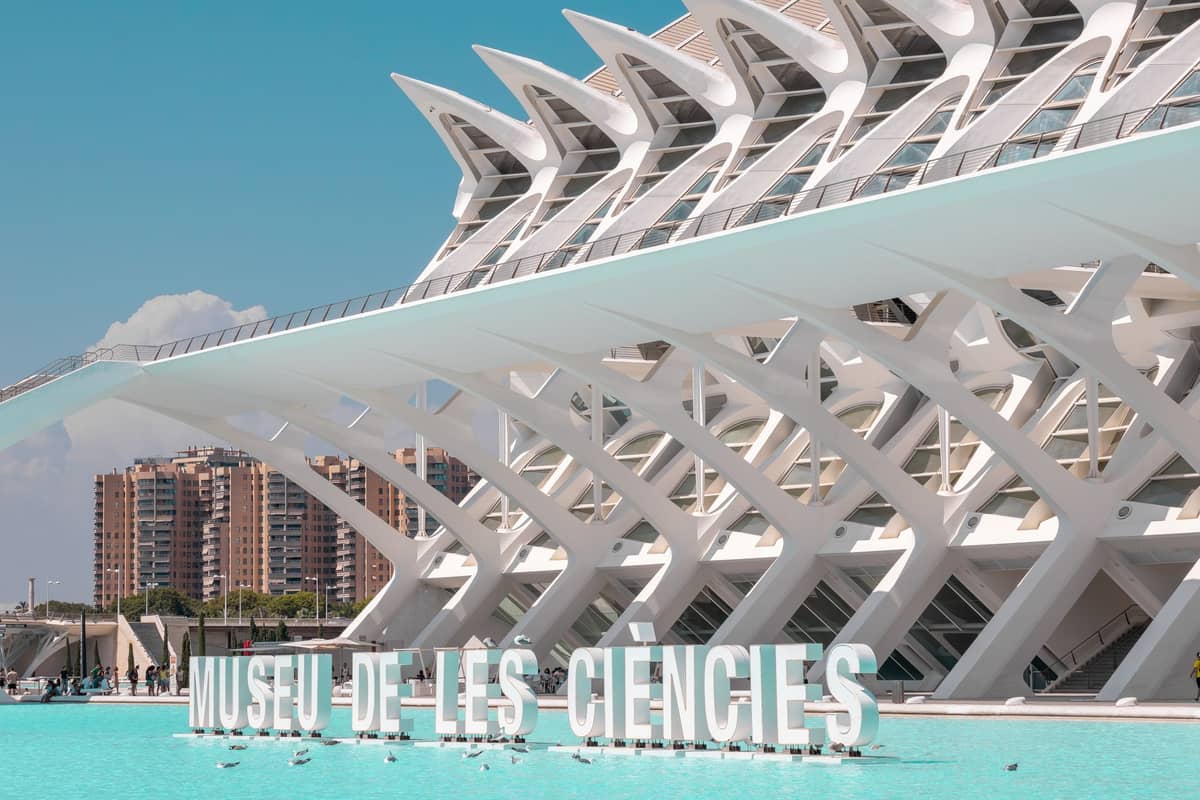 Vista esterna del Museu de les Ciències Príncep Felip a Valencia, con la sua iconica struttura bianca a scheletro e il cartello del nome riflesso nell'acqua azzurra.