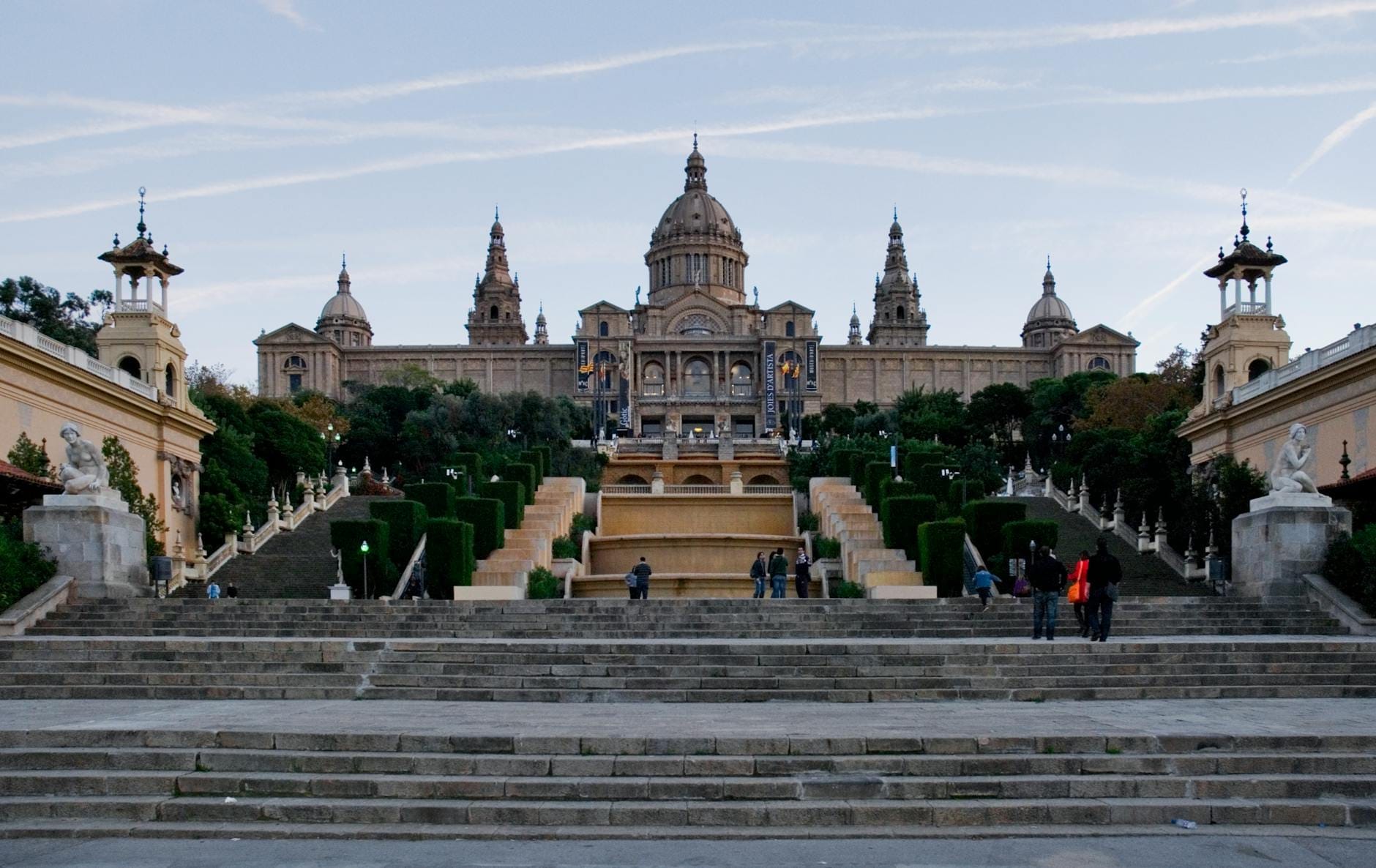Wide view of the Museu Nacional d'Art de Catalunya atop Montjuïc hill, framed by grand stairways, gardens, and visitors exploring the approach.