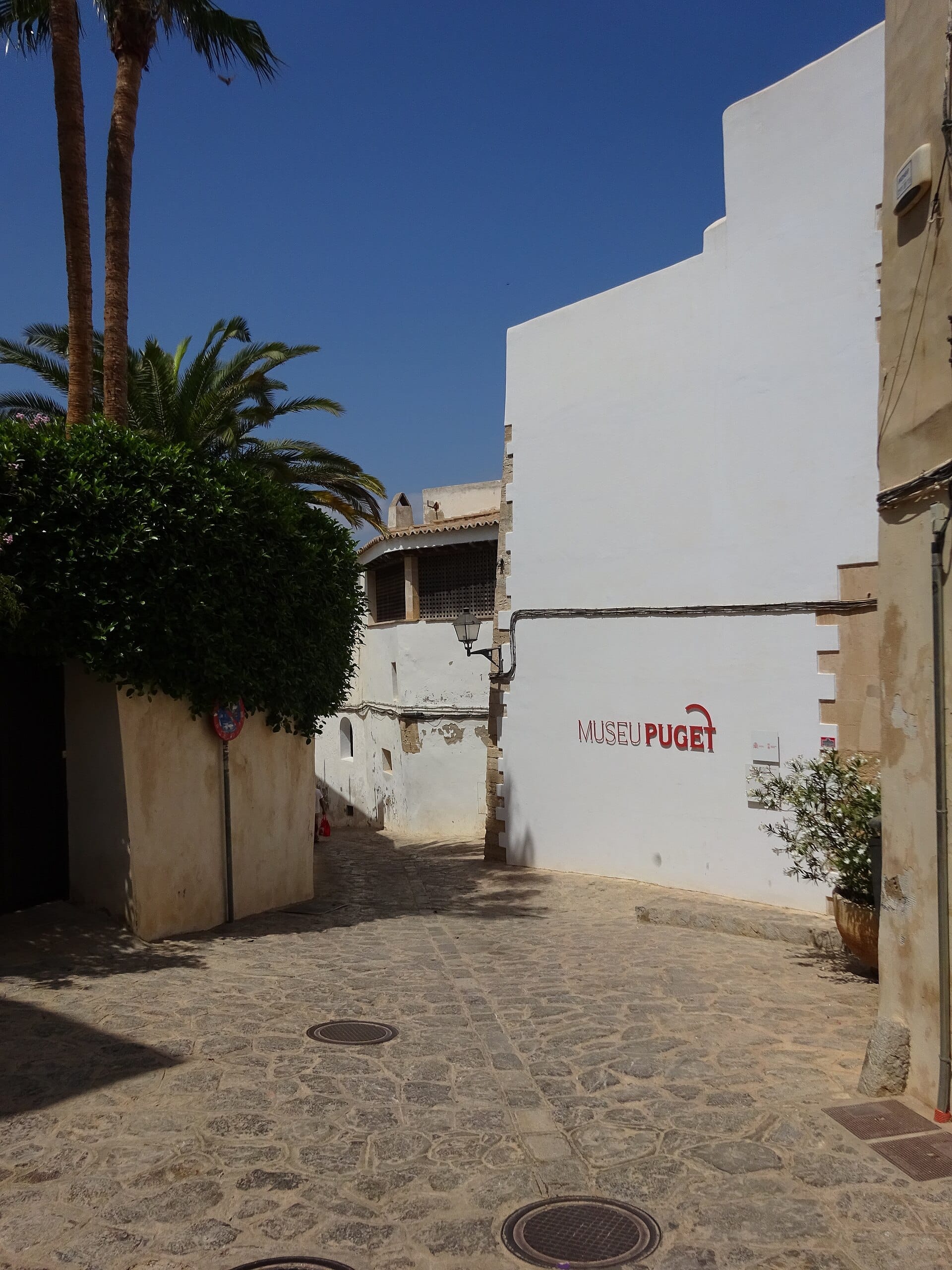 Stretto vicolo in pietra che conduce alla facciata imbiancata del Museu Puget a Ibiza, con palme e un cielo azzurro brillante.