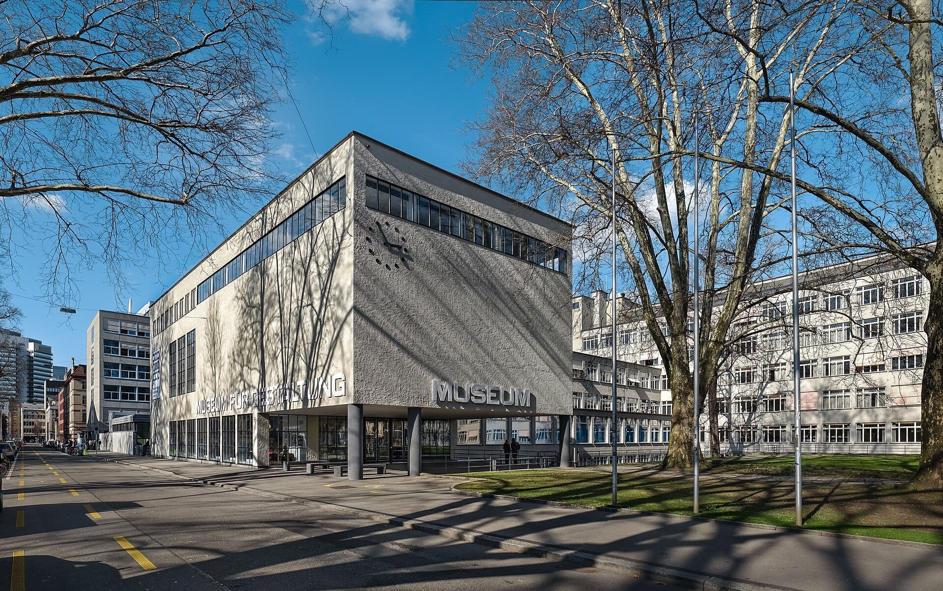 ด้านหน้าอาคารแบบโมเดิร์นนิสต์ของ Museum für Gestaltung Zürich พร้อมนาฬิกาขนาดใหญ่ หน้าต่างกระจก และกิ่งไม้ที่ไม่มีใบภายใต้ท้องฟ้าสีฟ้าแจ่มใส