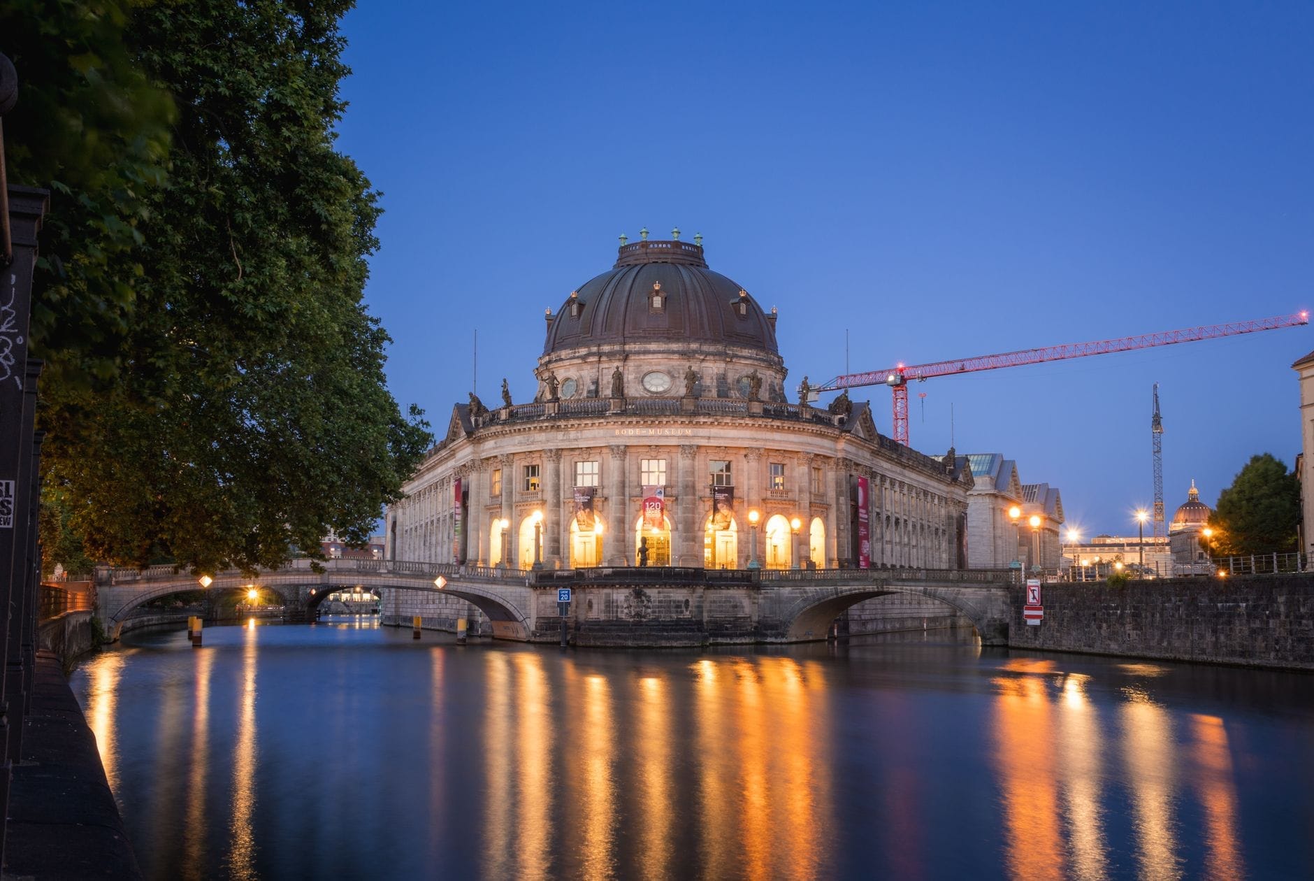 Vista serale del Bode Museum di Berlino sull'Isola dei Musei, splendidamente illuminato e rispecchiato nelle acque della Sprea sotto un cielo blu sereno.