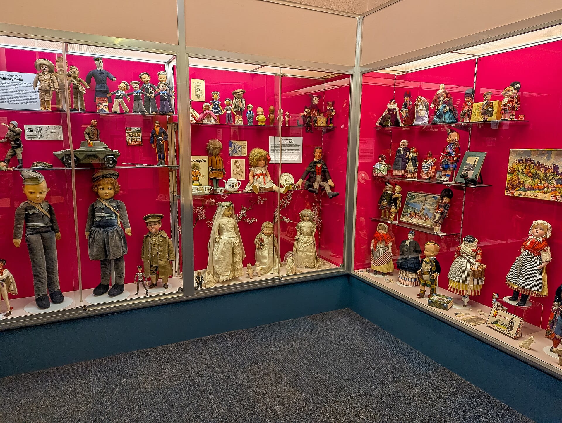 Breiter Blick auf Glasvitrinen mit Vintage-Puppen und Spielzeug im Museum of Childhood Edinburgh vor leuchtend roten Hintergründen.