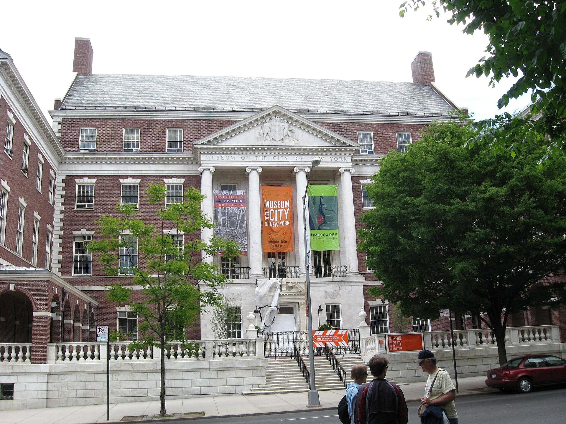 Facciata in mattoni del Museum of the City of New York con striscioni appesi tra colonne bianche, alberi e alcune persone sul marciapiede.