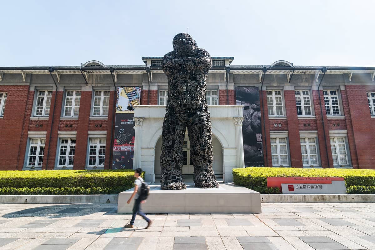 Die rotgeklinkerte Fassade des Museum of Contemporary Art Taipei mit Säulen, Bannern und einer großen modernen Skulptur vor dem Eingang.