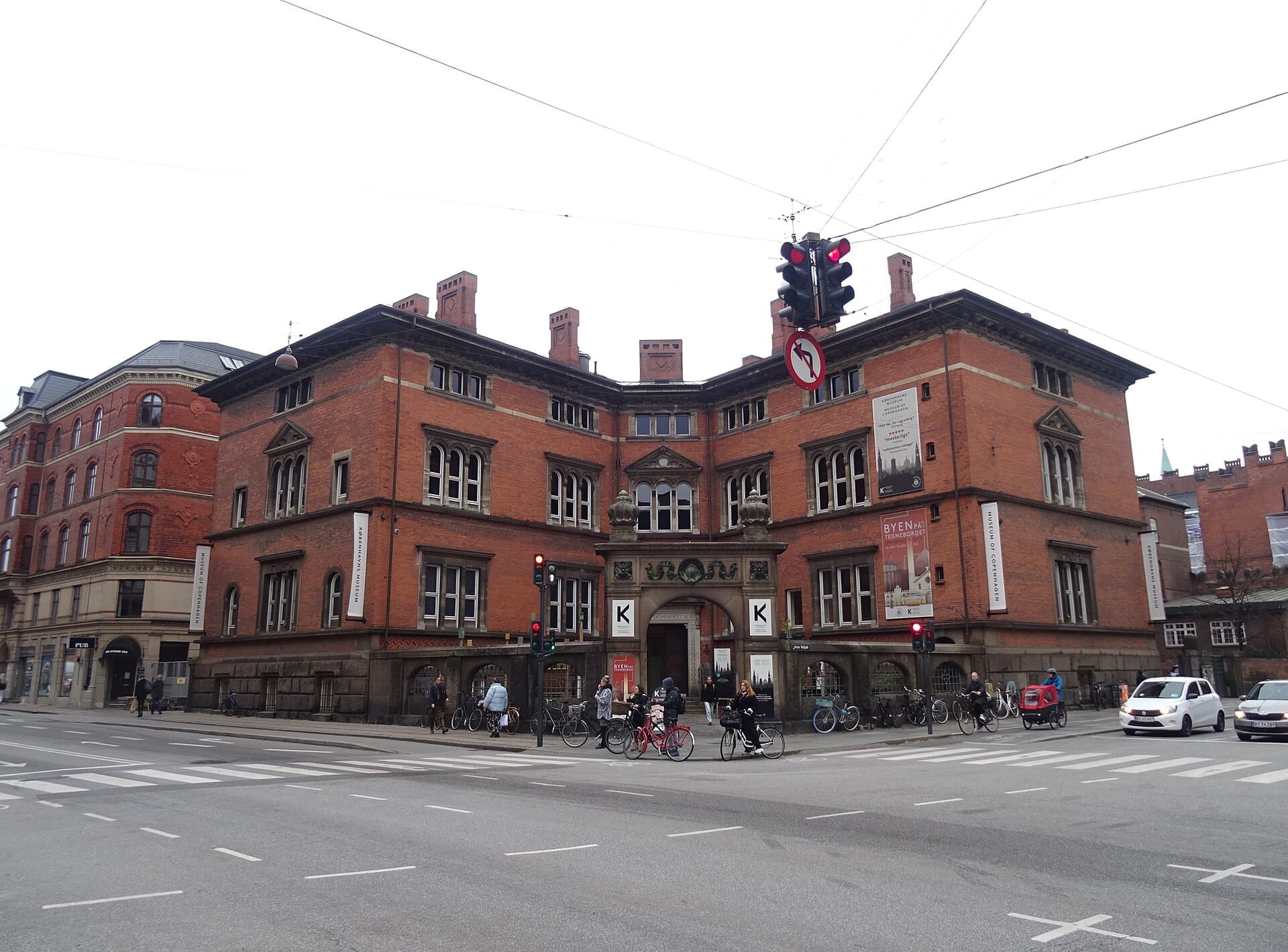 Edificio in mattoni rossi del Museum of Copenhagen in una giornata nuvolosa, con ciclisti, pedoni e auto all'affollato incrocio antistante.