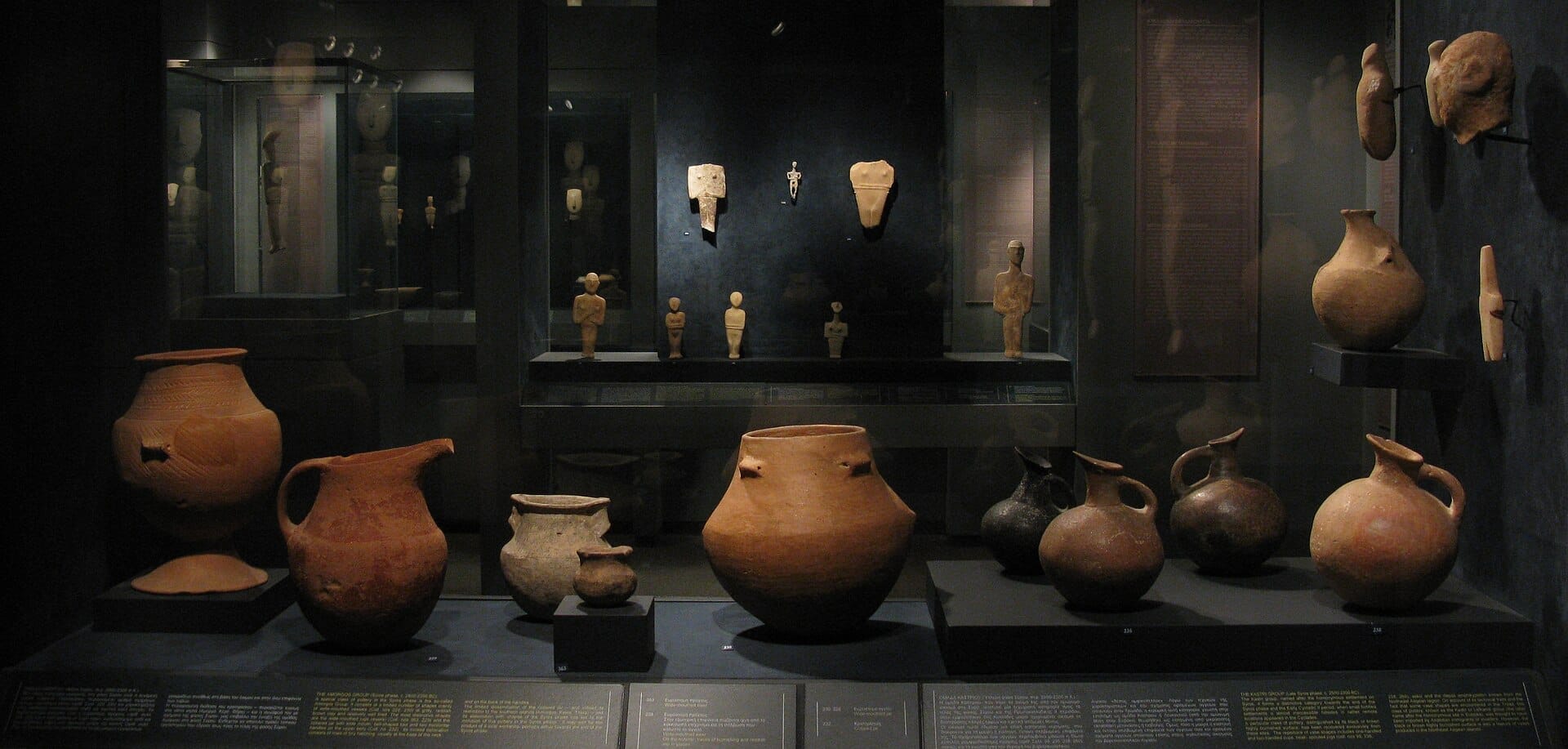 Esposizione al Museo dell'Arte Cicladica con antichi vasi in terracotta e figurine cicladiche esposti in vetrine fiocamente illuminate.