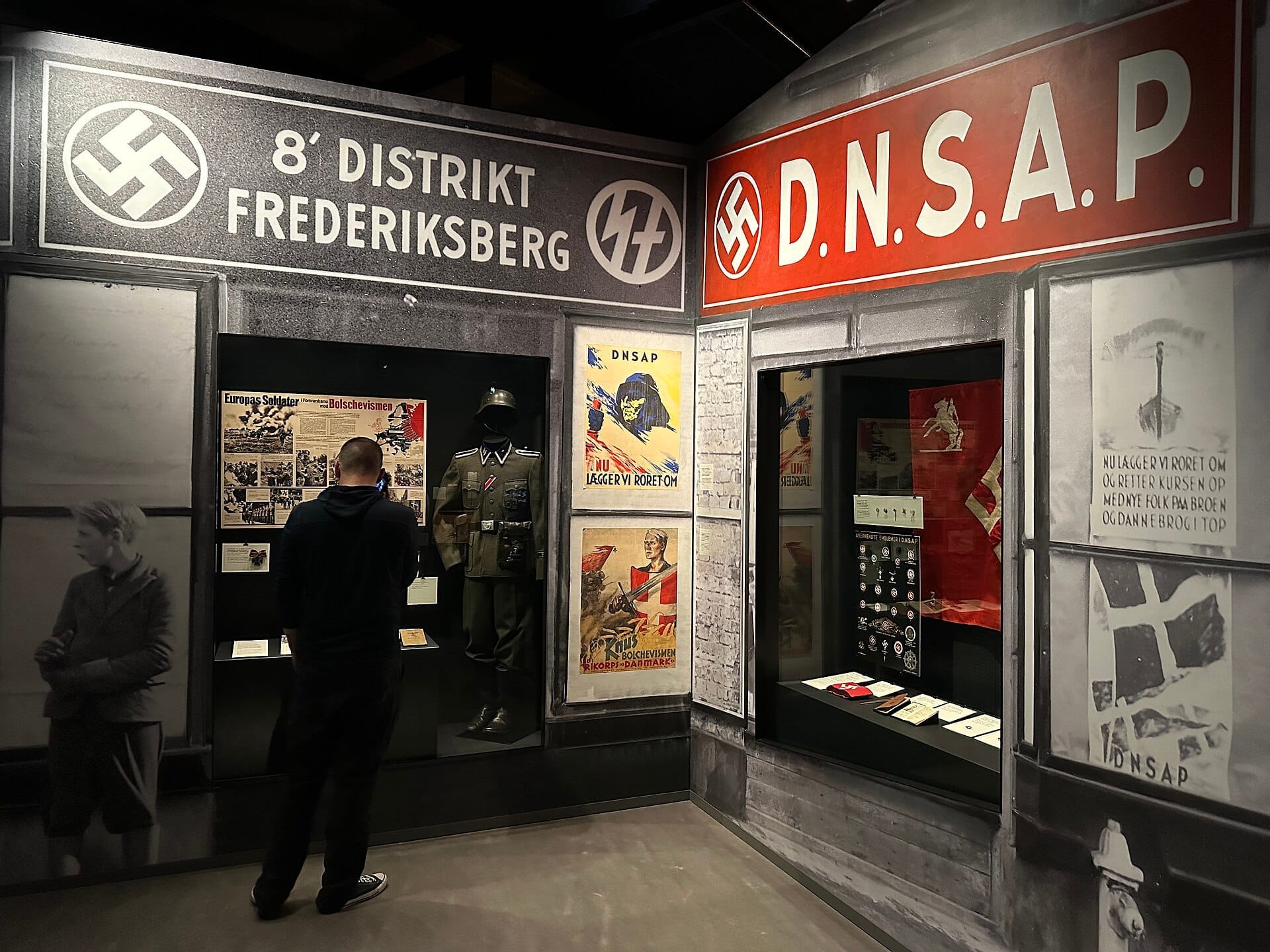 Un visitatore esamina reperti della Seconda Guerra Mondiale e cimeli nazisti al Museum of Danish Resistance di Copenaghen, circondato da manifesti storici e uniformi.