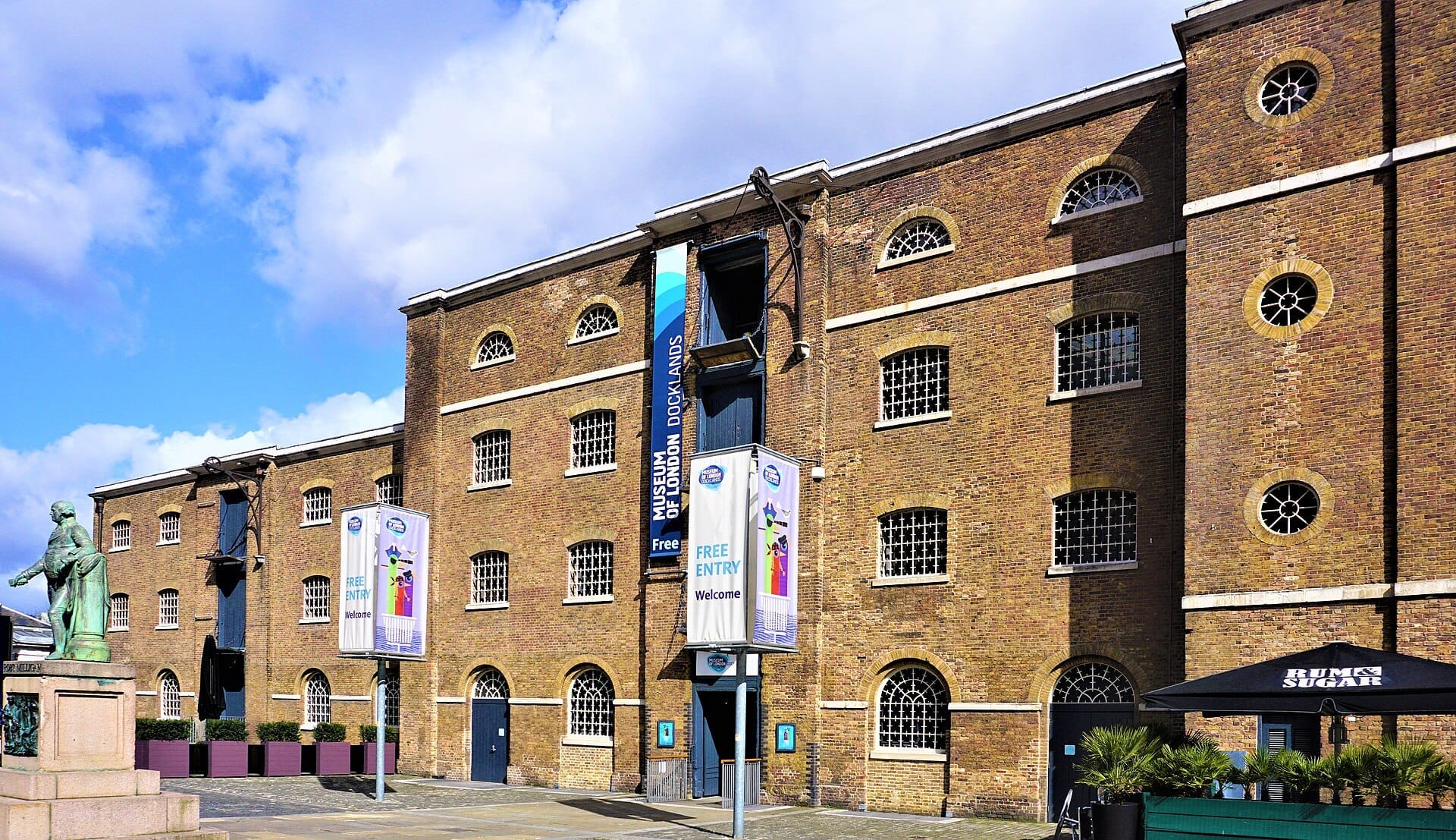Esterno del London Museum Docklands, uno storico magazzino in mattoni con finestre ad arco, statua e striscioni per visitatori sotto un cielo azzurro brillante.