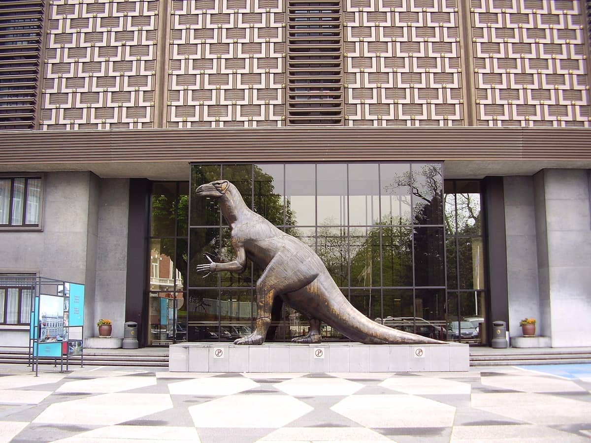 Uma grande estátua de dinossauro em bronze diante do Museum of Natural Sciences Brussels, com arquitetura moderna de vidro e pedra geométrica ao fundo.
