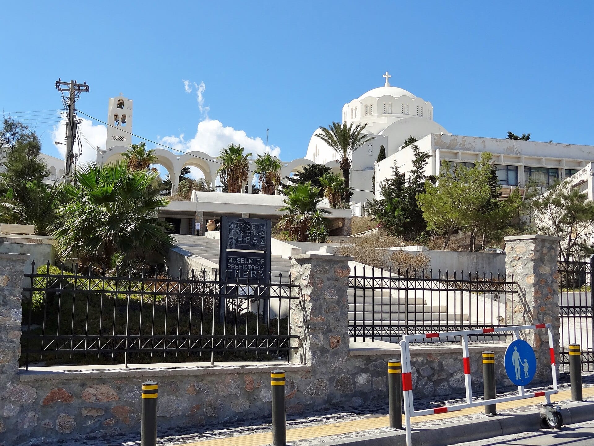 วิวถนนของพิพิธภัณฑ์ยุคก่อนประวัติศาสตร์ Thera ใน Fira, Santorini พร้อมด้านหน้าอาคารสีขาวทันสมัย ต้นปาล์ม และป้ายทางเข้าในวันที่แดดจัด