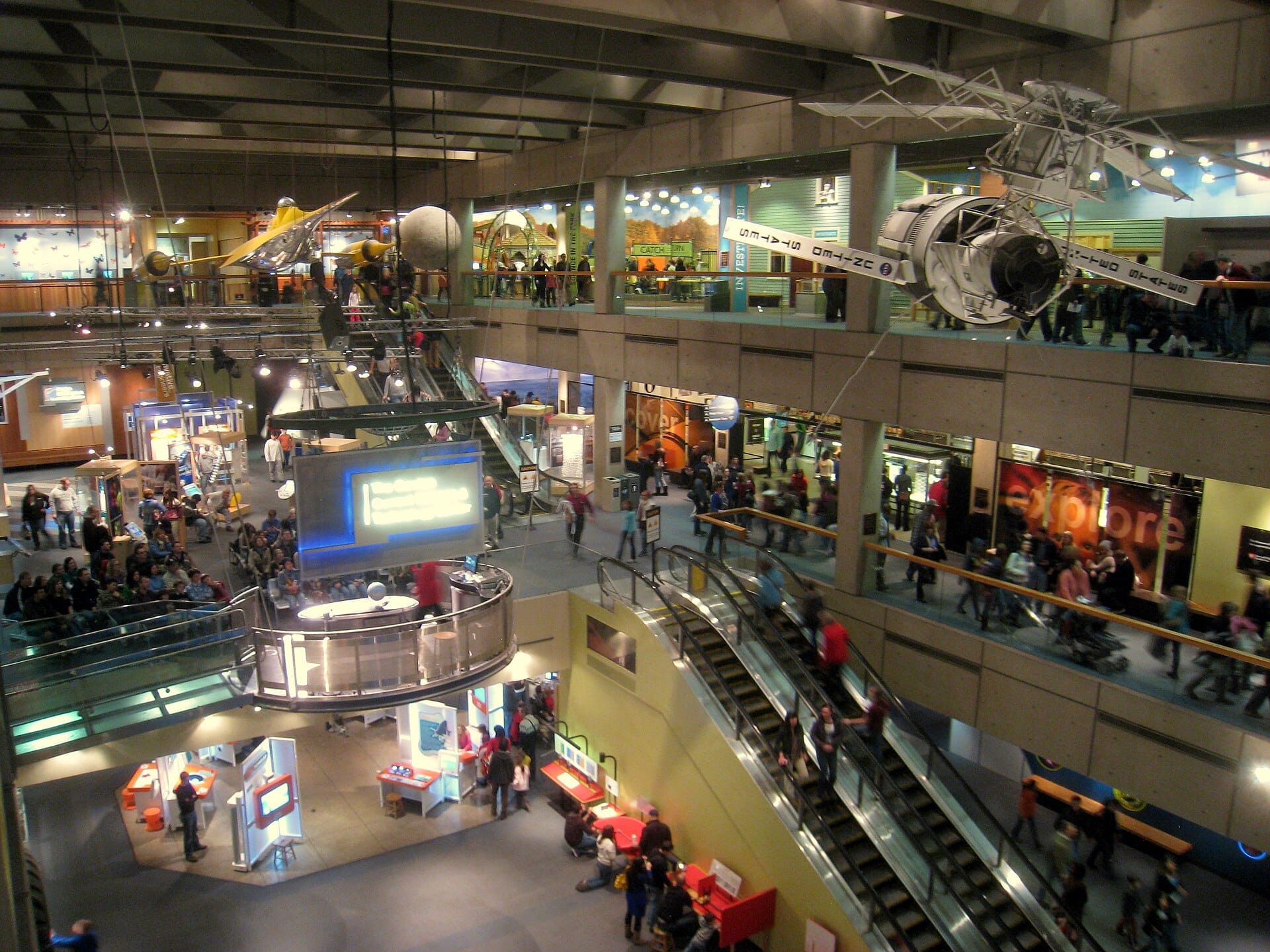 Vista ampia dell'interno del Museum of Science di Boston, con esposizioni interattive, scale mobili e modelli di veicoli spaziali appesi, mentre i visitatori esplorano più livelli.