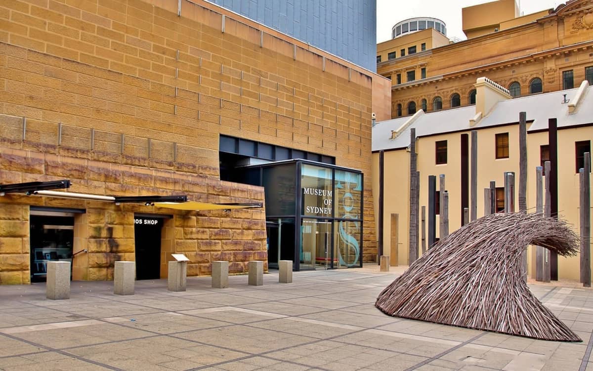 Ingresso al Museum of Sydney, con pareti in arenaria, porte in vetro moderne e una grande scultura a forma di onda nella piazza esterna.