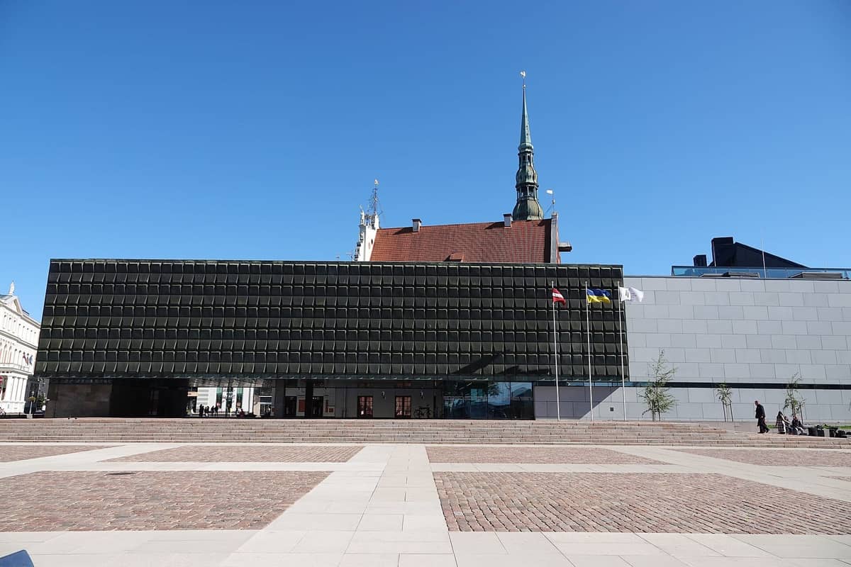 Die Außenfassade des Okkupationsmuseums Lettland in Riga mit seiner markanten modernen, rechteckigen Fassade, Flaggen und einem großen öffentlichen Platz im Vordergrund.