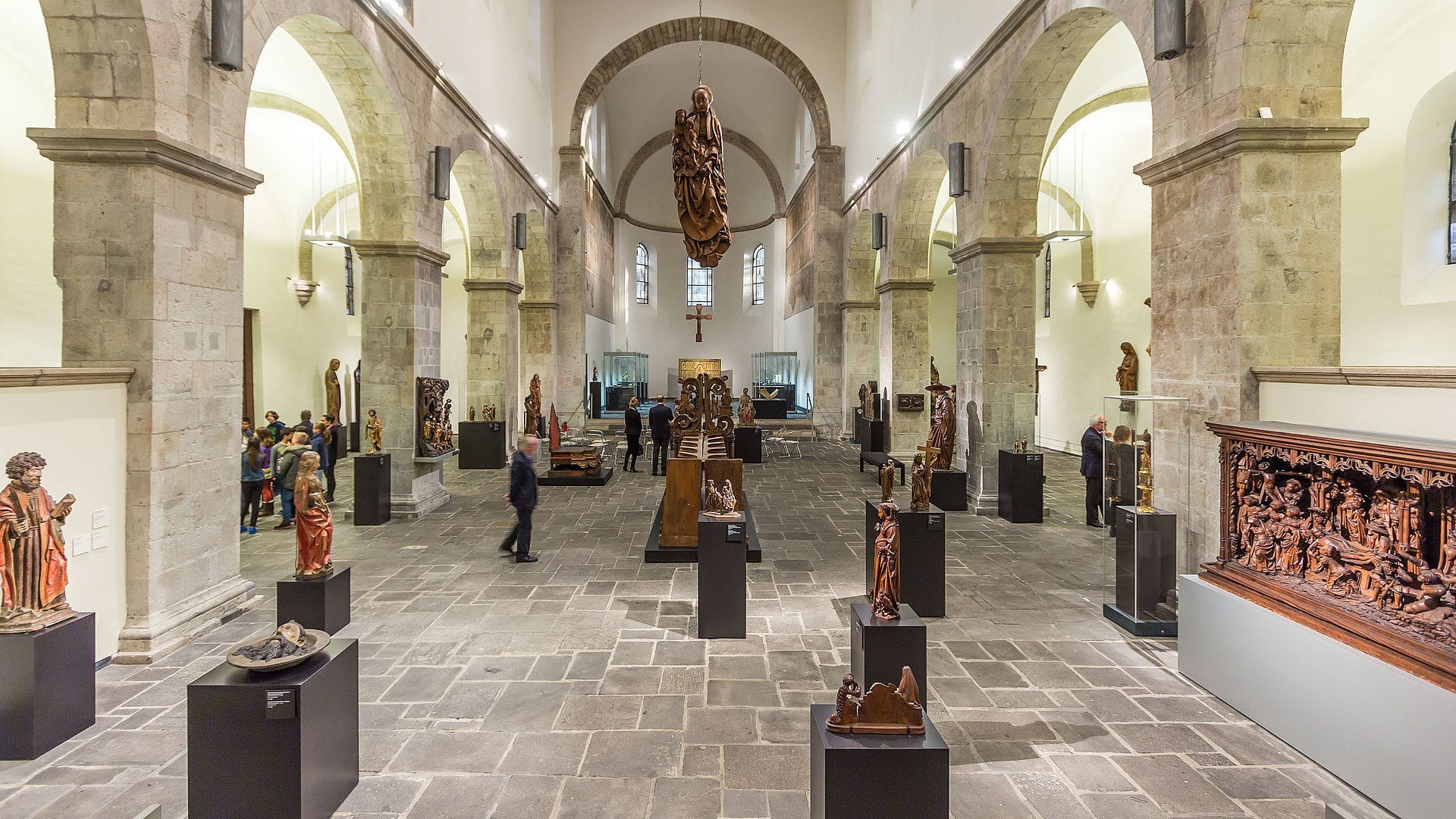 มุมมองภายในของ Museum Schnütgen พร้อมประติมากรรมและงานแกะสลักคริสเตียนยุคกลางที่จัดแสดงระหว่างซุ้มโค้งหินโรมาเนสก์ภายใต้แสงสว่างจ้า