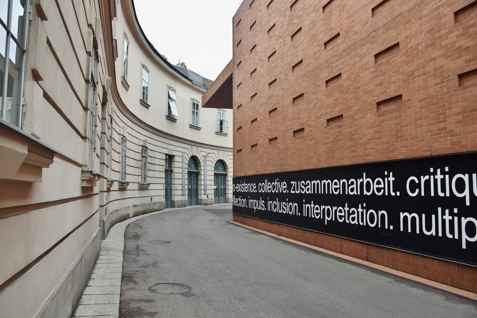 Geschwungene Straße zwischen historischen und modernen Gebäuden des MuseumsQuartiers in Wien, mit zeitgenössischer Textkunst auf einer Backsteinmauer und keinen Menschen im Bild.