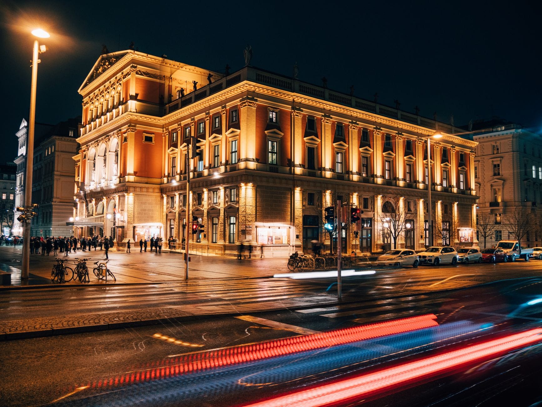 Nächtlicher Blick auf das illuminierte Musikverein in Wien mit seiner prachtvollen neoklassizistischen Fassade, belebtem Straßenleben und Lichtspuren von Autos im Vordergrund.