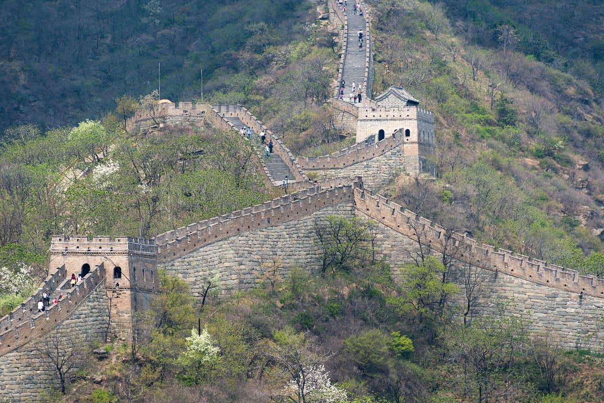 Ein weiter Blick auf den Mutianyu-Abschnitt der Chinesischen Mauer mit mehreren Wachtürmen, steinernen Stufen und grünen, bewaldeten Hügeln rund um die Mauer, auf der Besucher entlangspazieren.