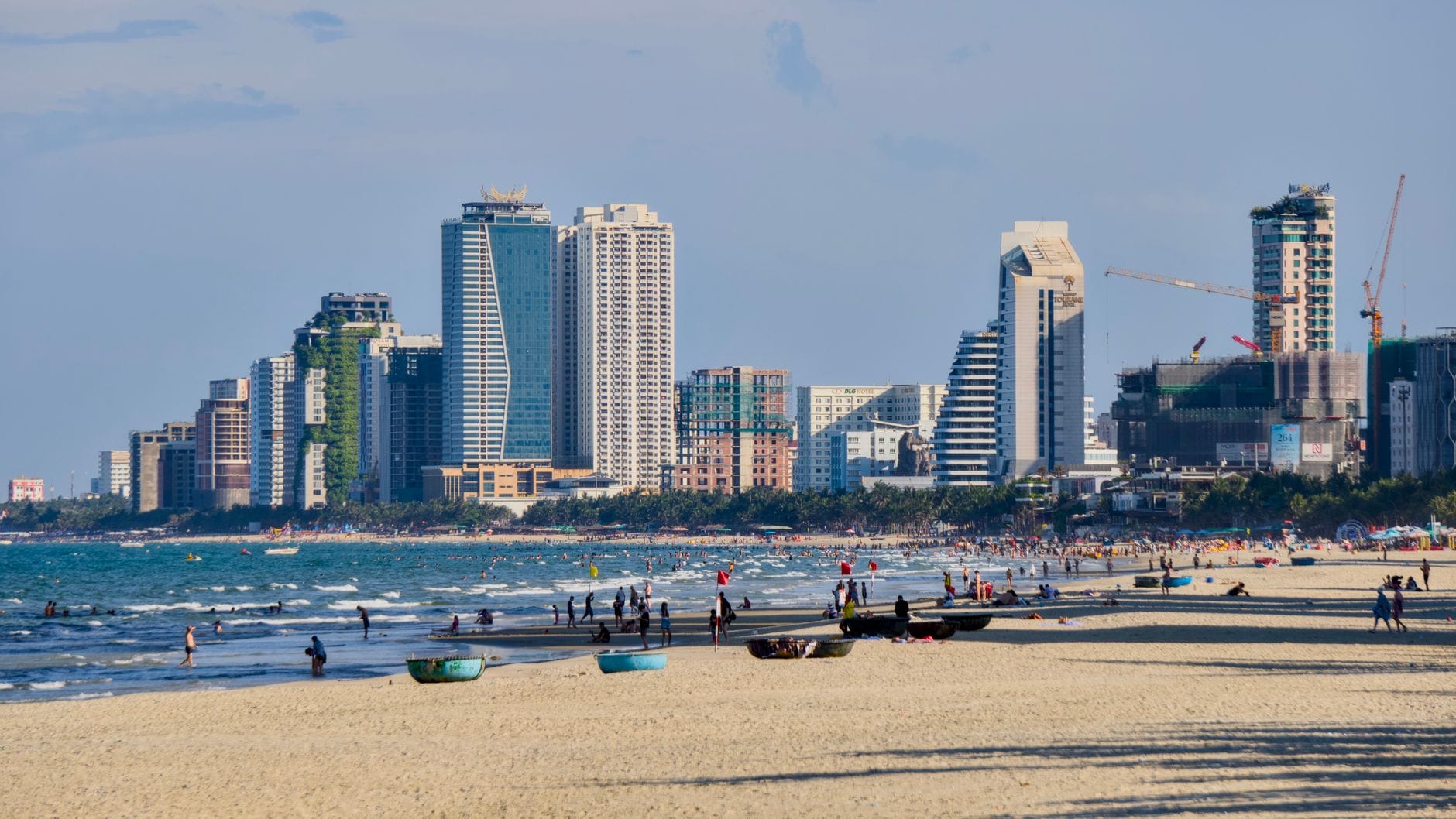 ภาพทิวทัศน์หาดมายเค คนเดินเล่น เรือประมง เมืองดานังอยู่พื้นหลังกับตึกสูง ภายใต้ท้องฟ้าสีฟ้า