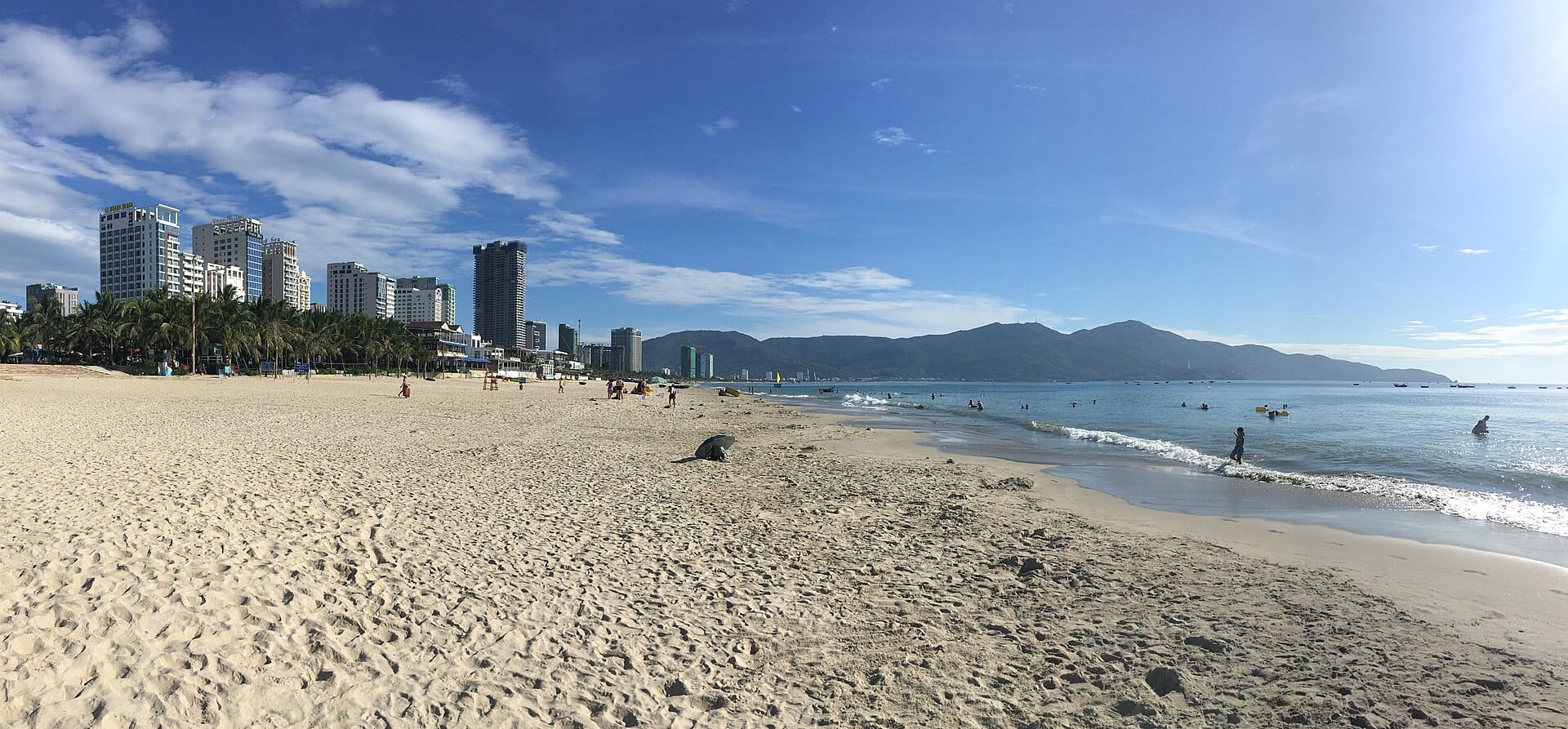 หาดมายเคผืนทรายกว้าง มีคนว่ายน้ำ ปาล์ม และตึกระฟ้าของดานังใต้ท้องฟ้าใสกับวิวภูเขาไกลๆ