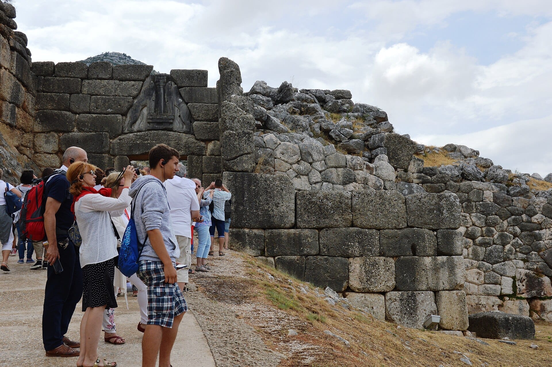 Visitatori in piedi vicino all'antica Porta dei Leoni, circondata da massicce mura ciclopiche nel sito archeologico di Micene sotto un cielo nuvoloso.