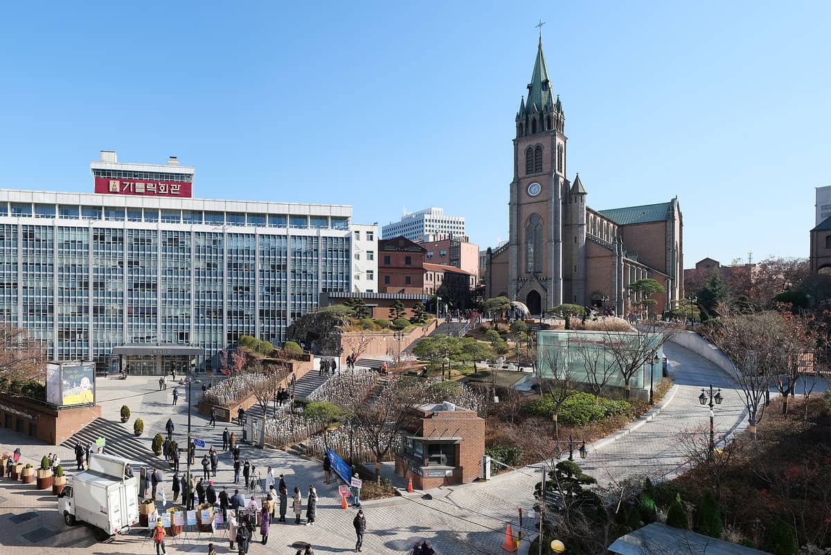 Vista ampla diurna da Catedral de Myeongdong no alto de uma colina acima de uma praça movimentada, com torre gótica, fachada em tijolos e prédios ao redor em Seul.