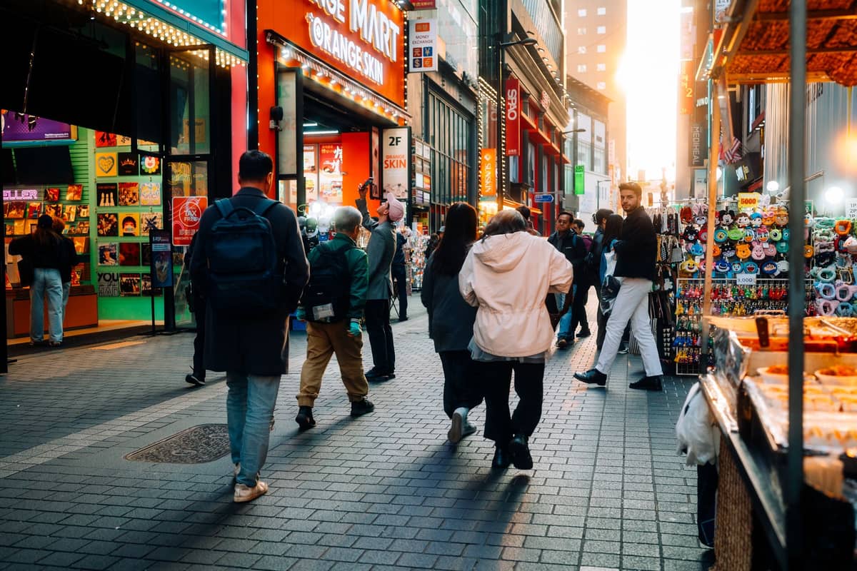 Rua movimentada de pedestres em Myeongdong, Seul, com lojas coloridas, barracas de comida de rua e pessoas caminhando à luz da tarde.