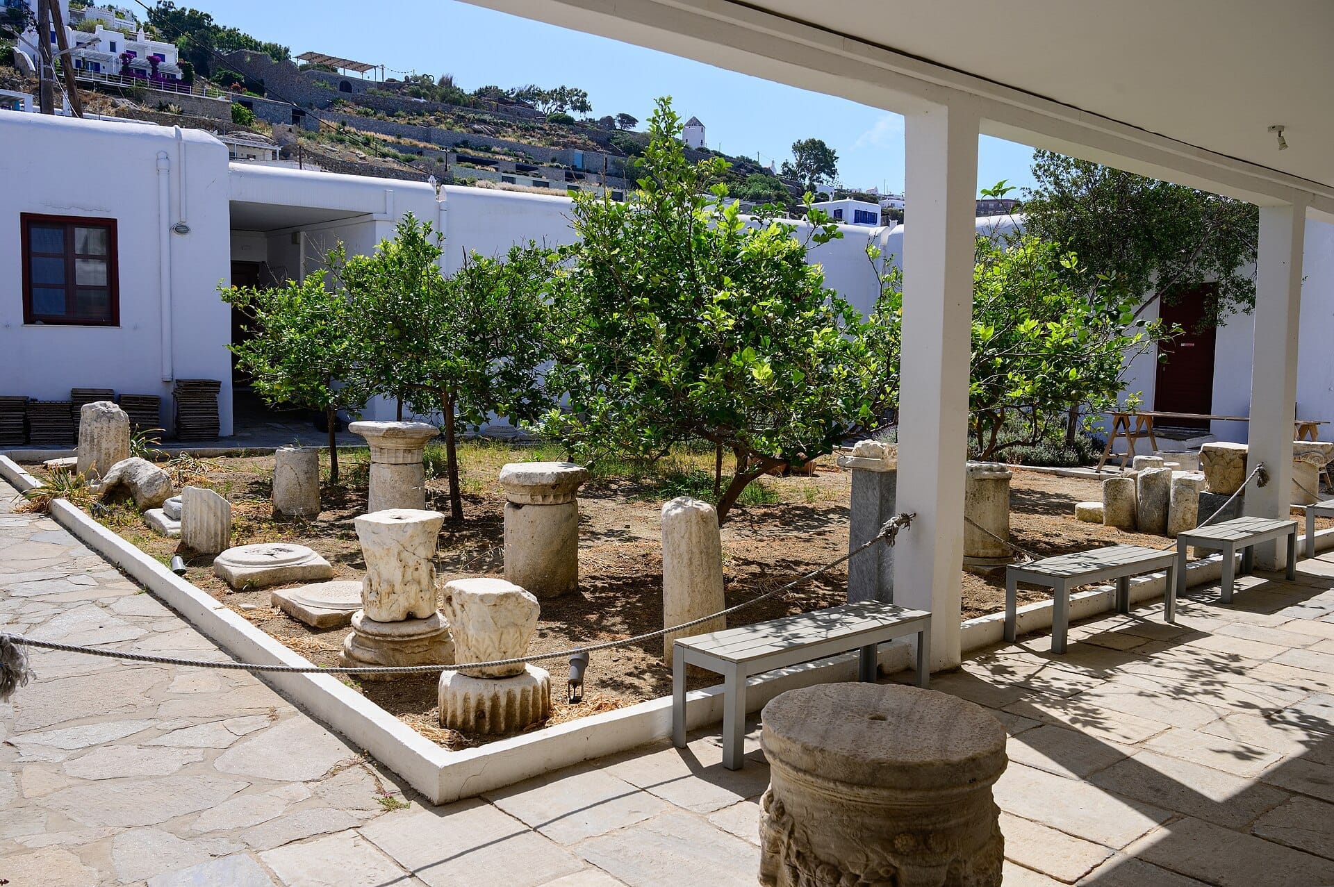 Antiguas columnas de mármol y artefactos de piedra expuestos en el patio soleado del Museo Arqueológico de Mykonos, rodeados de edificios encalados y vegetación.
