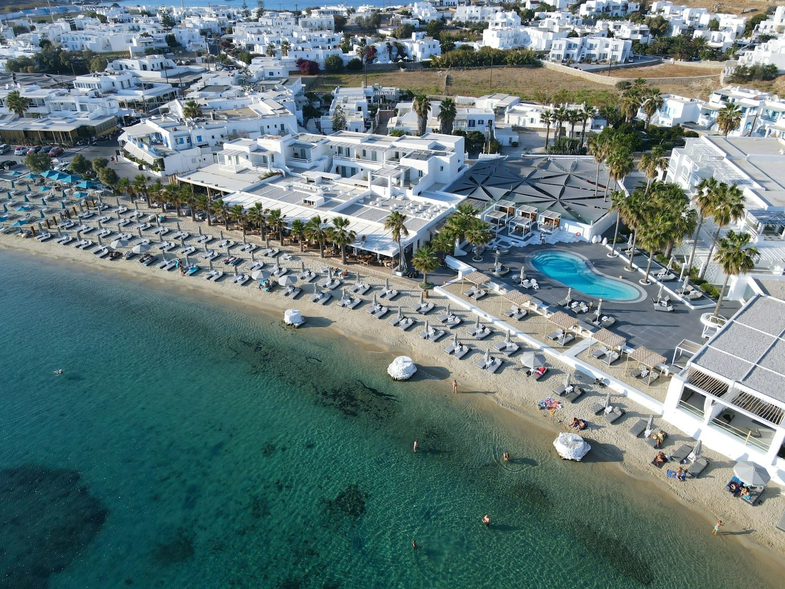 Vista aérea de un beach club en Mykonos con tumbonas, sombrillas, piscina y edificios encalados junto a un mar turquesa cristalino.
