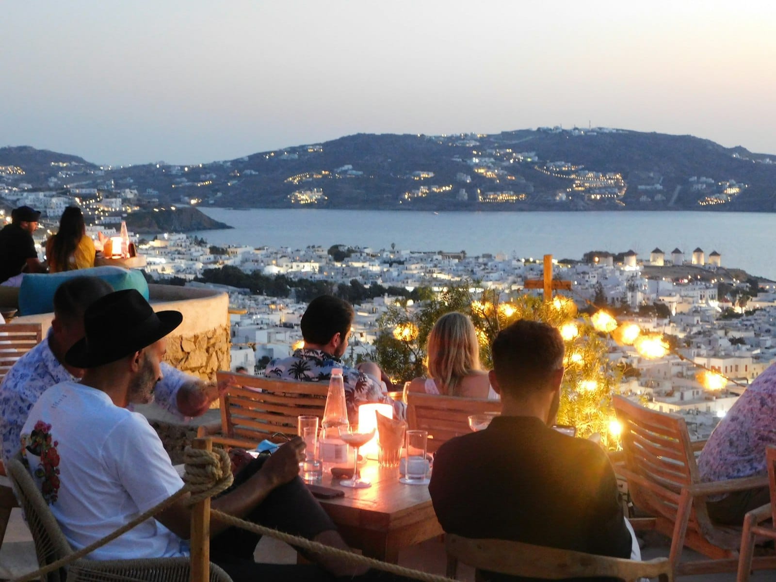 Personas disfrutando de bebidas al atardecer en una terraza exterior con vistas a Mykonos Town, el mar Egeo y los famosos molinos de viento.