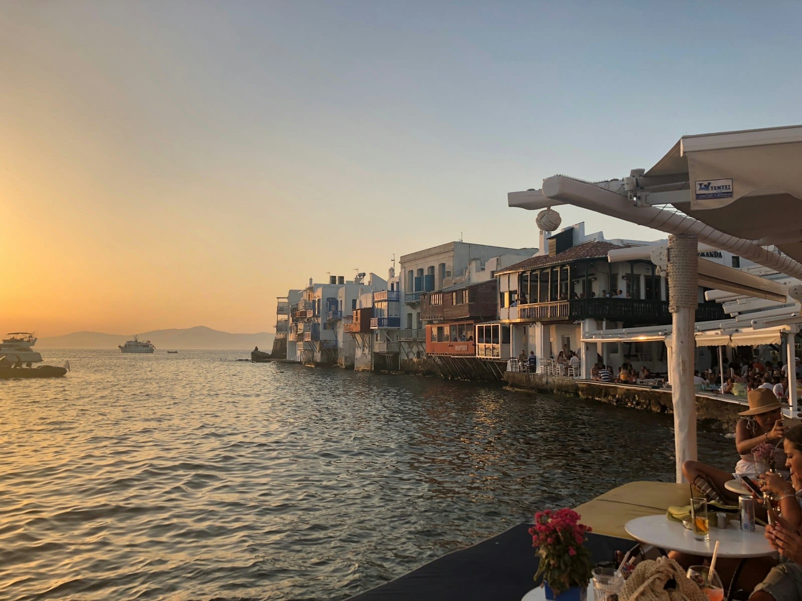 Restaurantes junto al mar y casas de colores de la Pequeña Venecia de Mykonos al atardecer, con mesas con vistas al agua y un romántico cielo anaranjado.