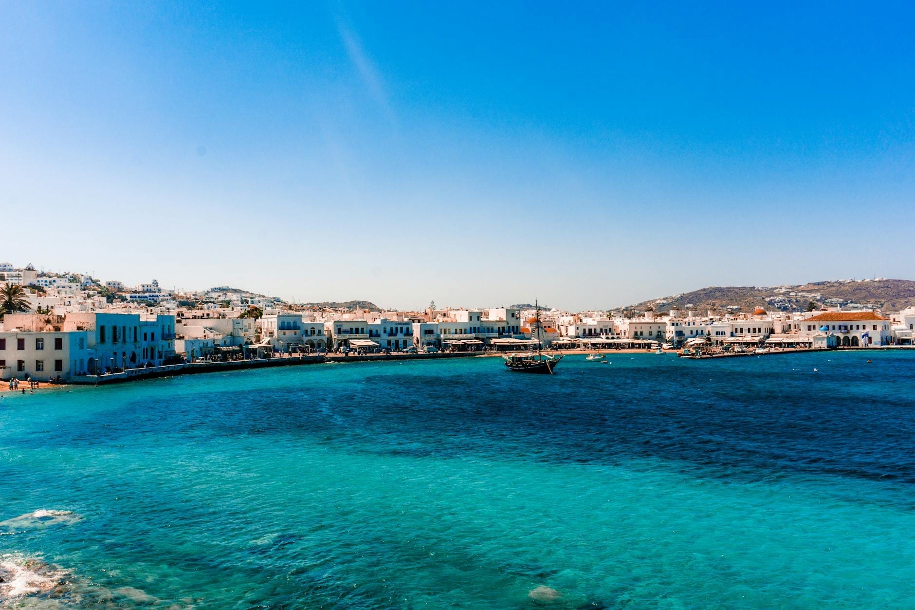 Mar Egeo de color azul brillante con barcos de lujo y el frente marítimo de la ciudad de Mykonos, edificios encalados y colinas al fondo bajo un cielo despejado.