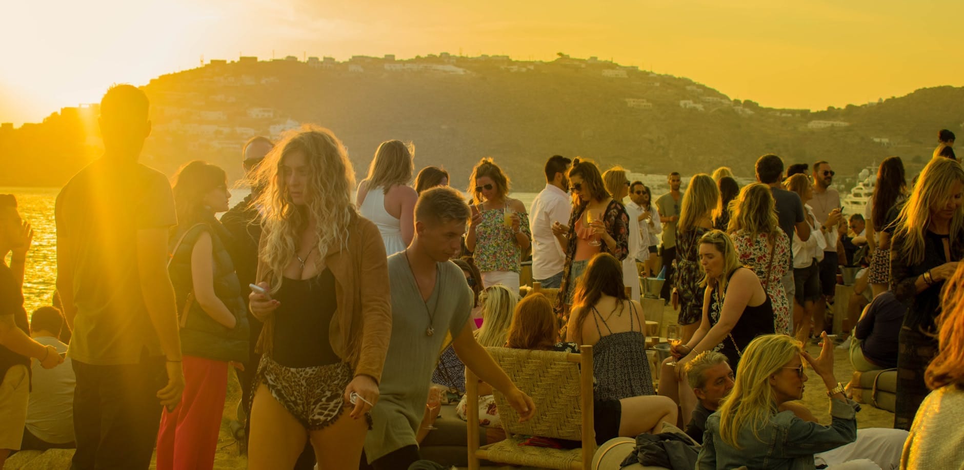 Multitud de personas elegantes disfrutando de un animado beach club al atardecer en Mykonos, sentadas y socializando cerca del mar con colinas al fondo.