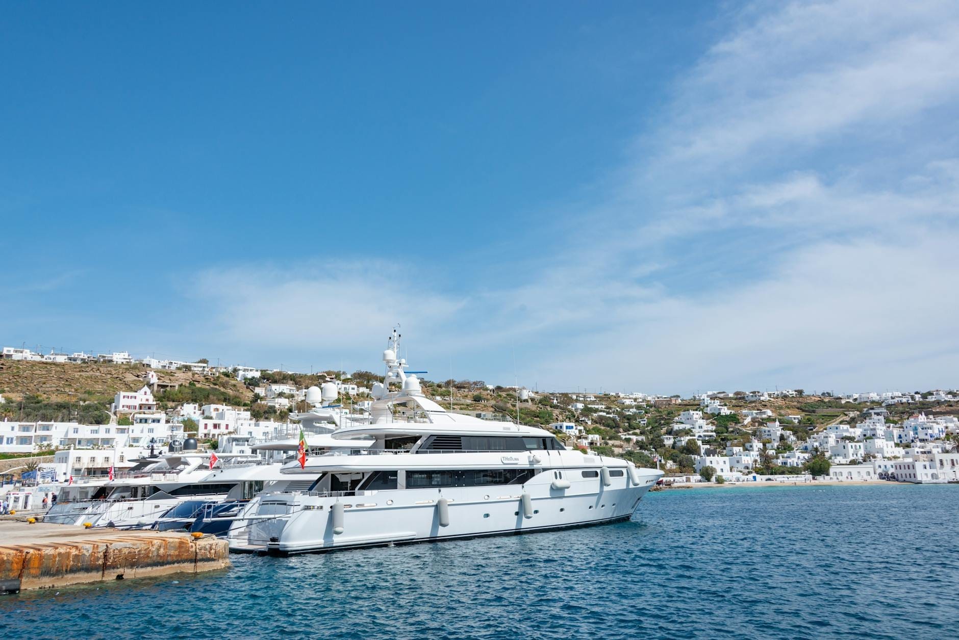 Gran yate de lujo atracado en el puerto de Mykonos con aguas cristalinas, edificios encalados y colinas bajo un cielo azul brillante.