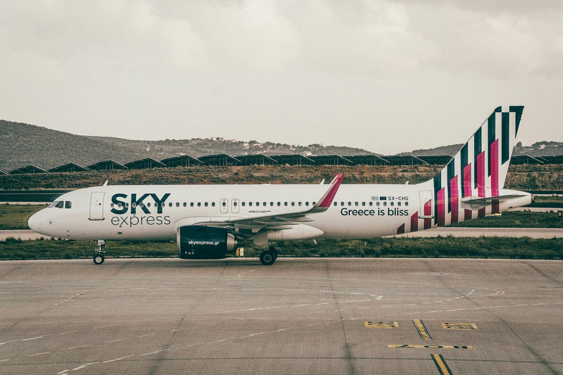 Avión de Sky Express con el eslogan 'Greece is bliss' estacionado en la pista de un aeropuerto de una isla griega, rodeado de colinas y cielo nublado.
