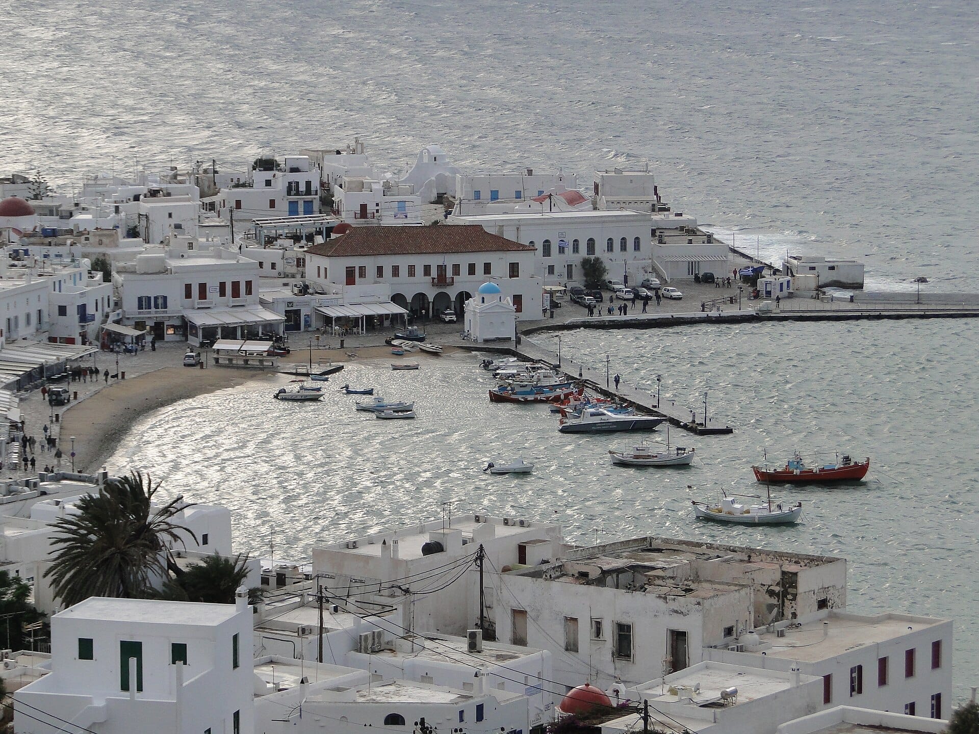 Vista panorámica del Puerto Antiguo de Mykonos con edificios encalados, pequeñas embarcaciones amarradas en el puerto y la suave curva de la playa del paseo marítimo.