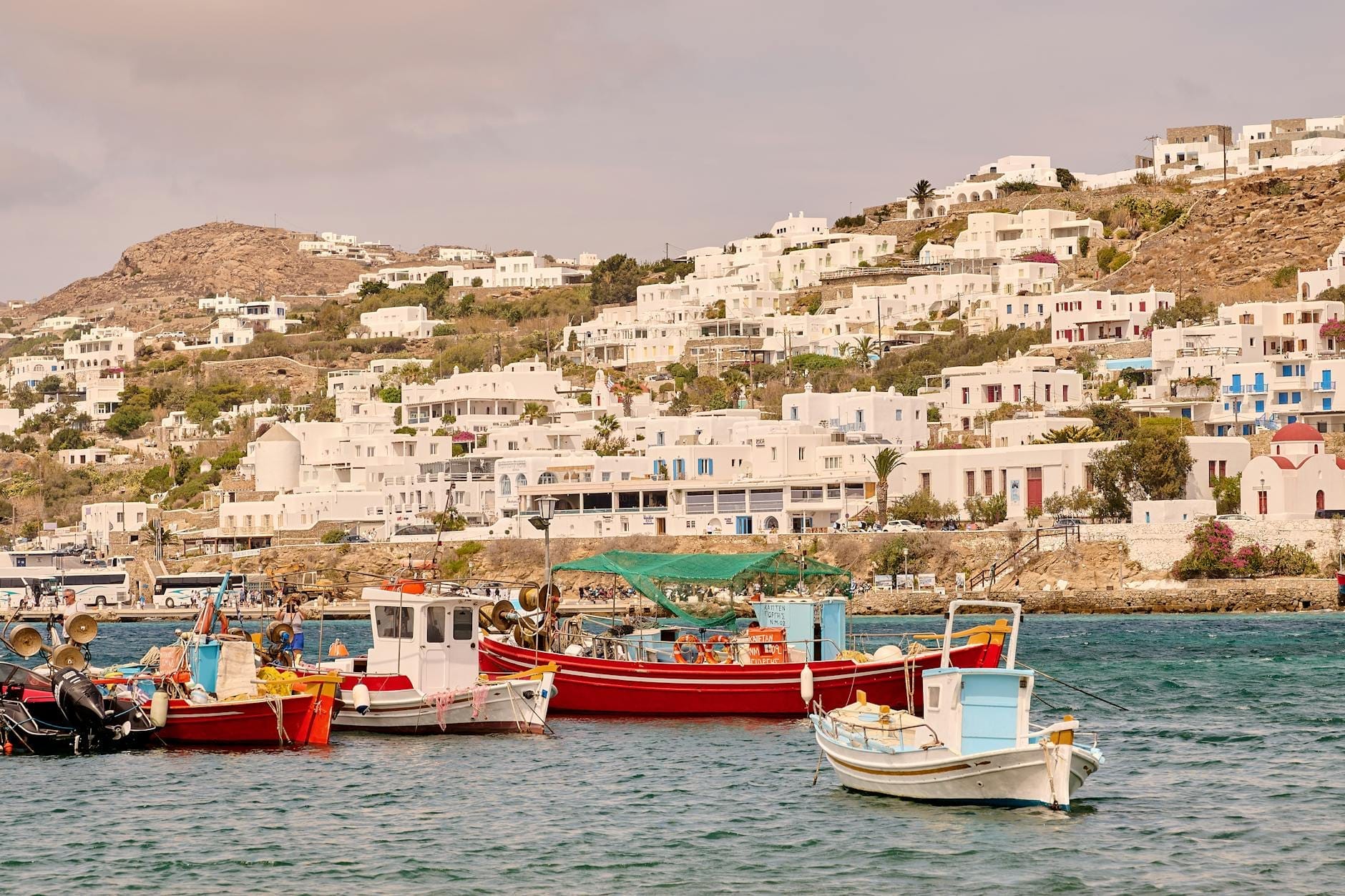 Barcos de pesca tradicionales amarrados en aguas turquesas con las casas encaladas de Mykonos visibles en la ladera al fondo.