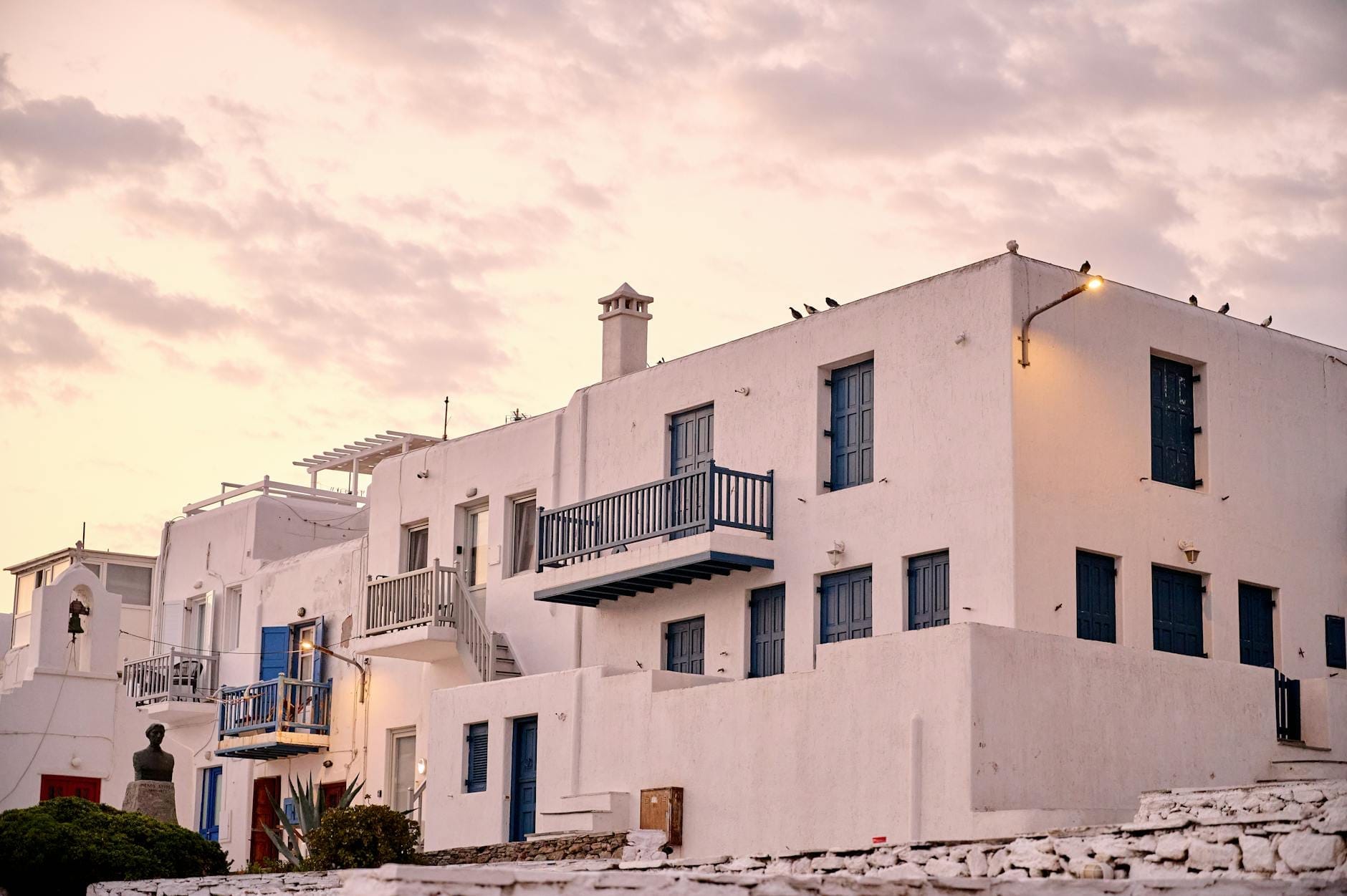 Edificios encalados con balcones y ventanas azules en Mykonos al atardecer, típicos de las opciones de alojamiento locales.