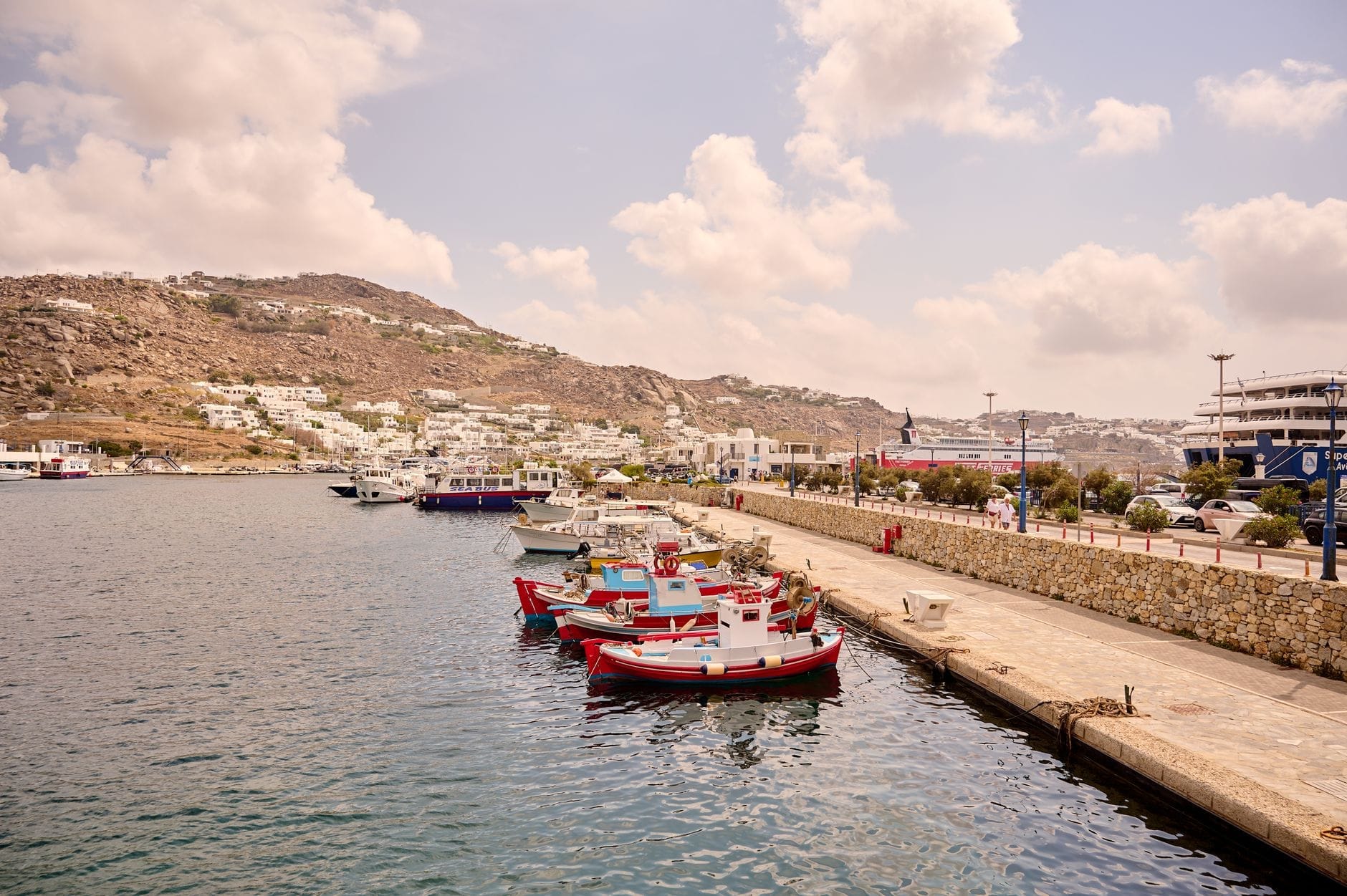 Varios barcos amarrados a lo largo del muelle en el Puerto Nuevo de Mykonos con edificios y colinas al fondo.