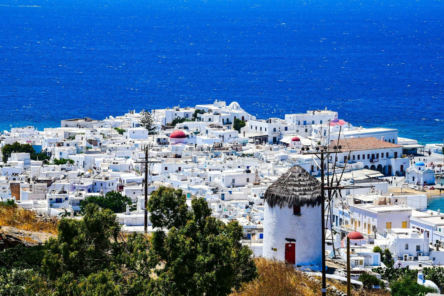 Vista panorámica de Mykonos Town con casas cicládicas blancas, un molino de viento y el mar azul profundo al fondo bajo un cielo despejado.
